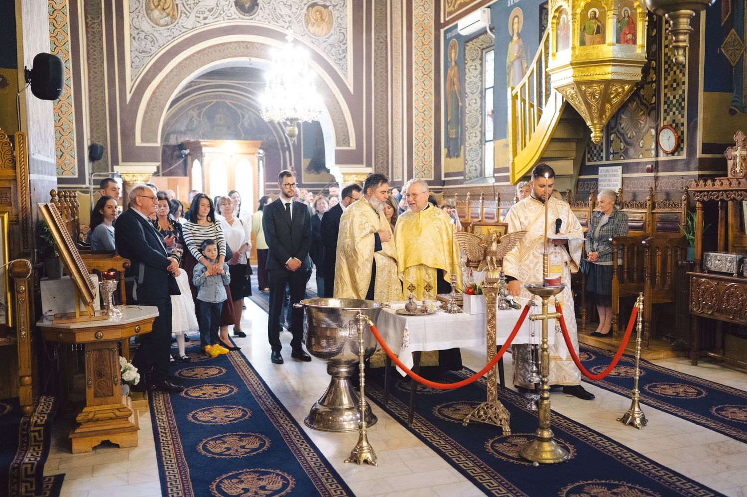 A religious ceremony takes place inside an ornate Orthodox church with golden decorations and a blue carpet runner.