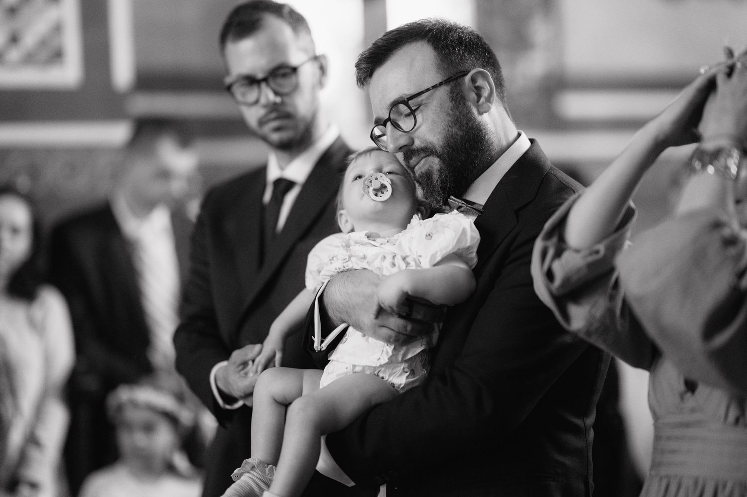 Black and white photo showing emotional moment as man in glasses holds a small baby at church gathering during ceremony.