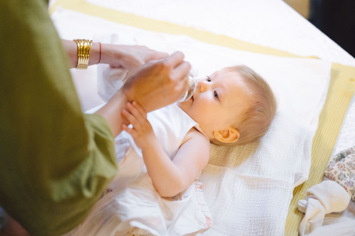 A baby lying on a white blanket reaches up while receiving medicine from a bottle at home.