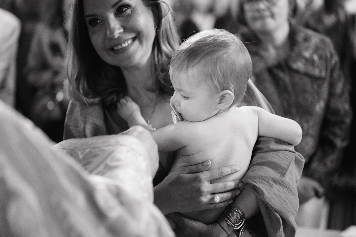 A tender black and white candid moment shows a baby being held at a gathering of people.