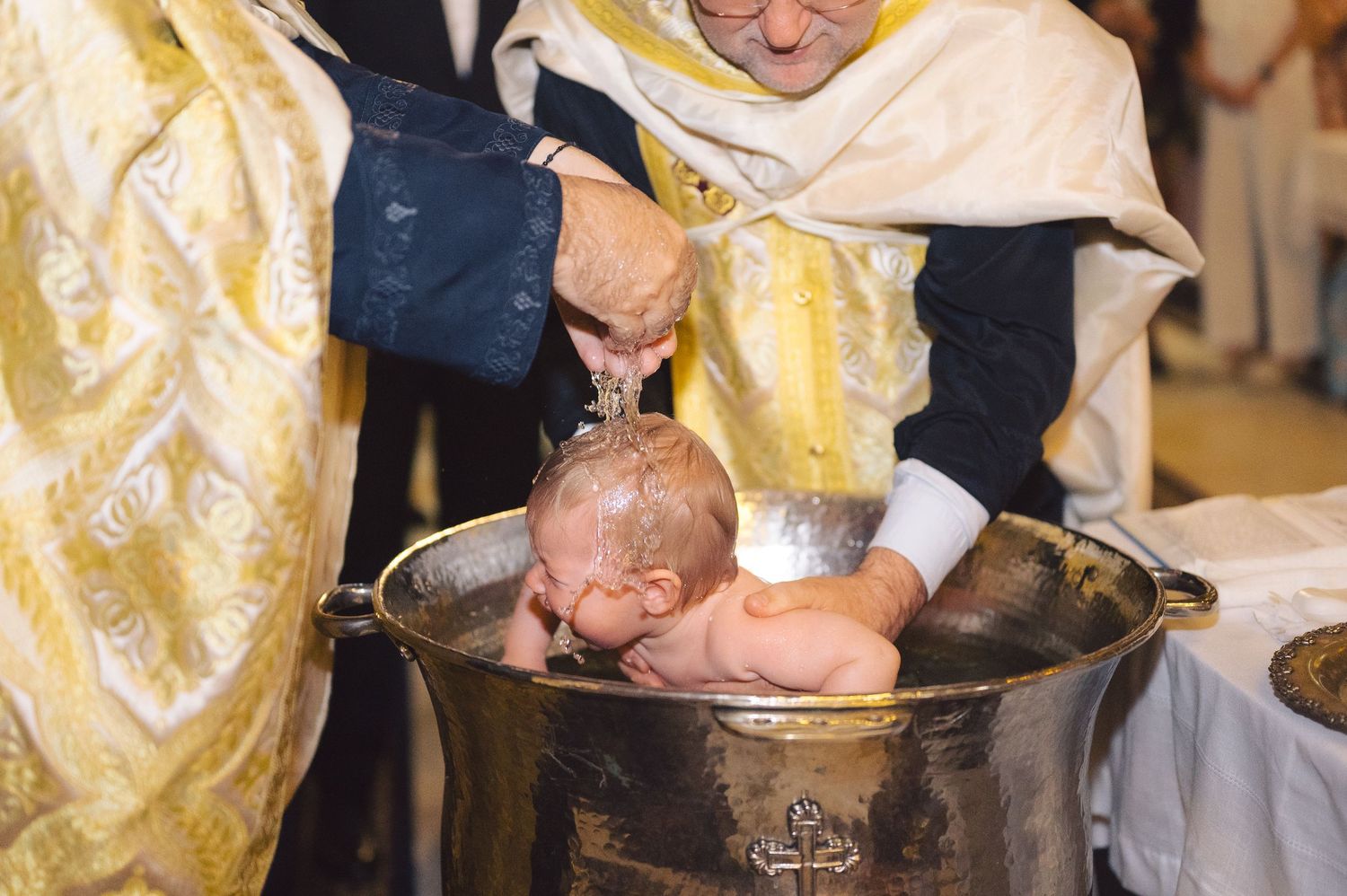 An Orthodox Christian baptism ceremony showing an infant being immersed in an ornate metal baptismal font.