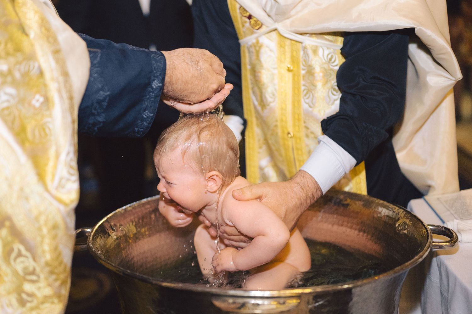 Orthodox Christian baptism ceremony showing infant being immersed in ornate brass basin during traditional ritual.