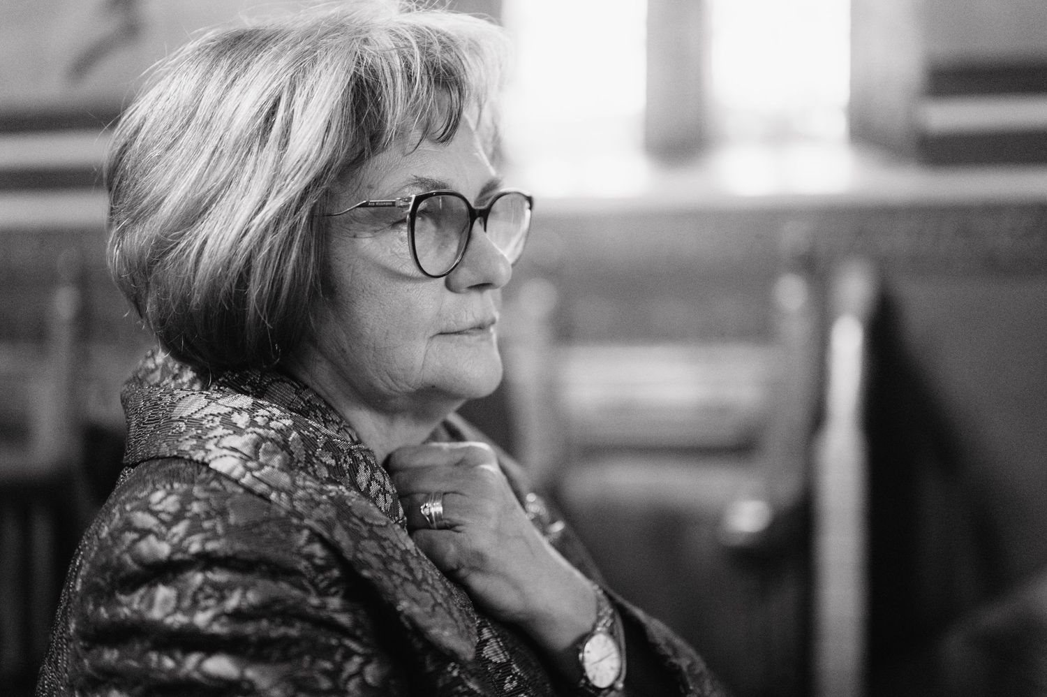 Black and white photo of a woman in glasses and patterned jacket gazing thoughtfully to the side with hand on chin.