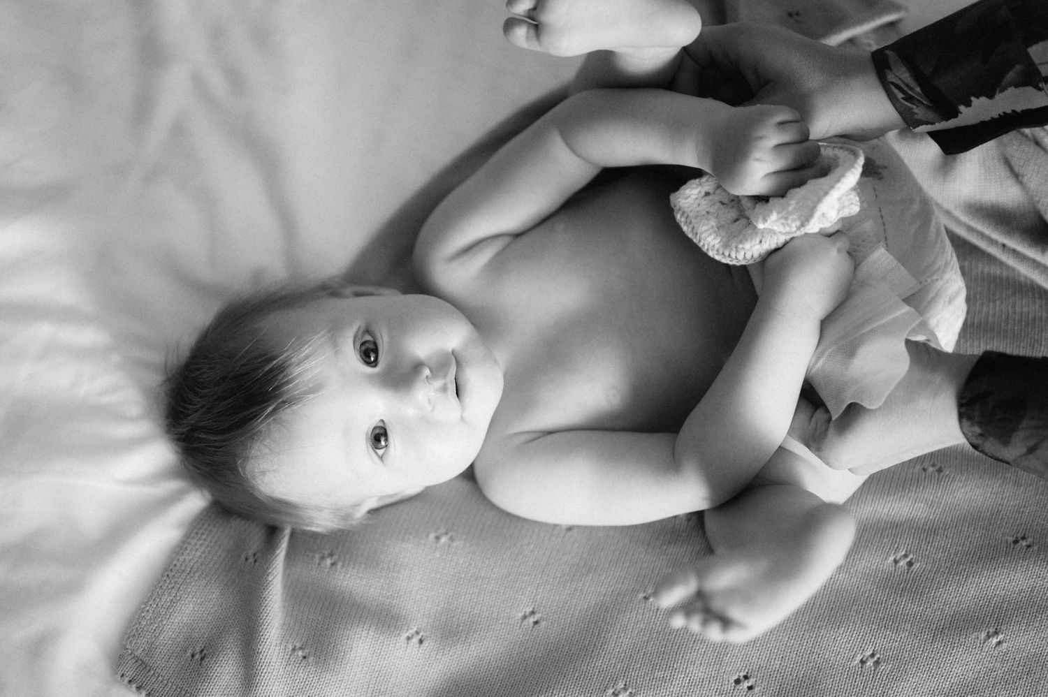 Black and white image of an infant lying on a textured blanket while holding a toy.
