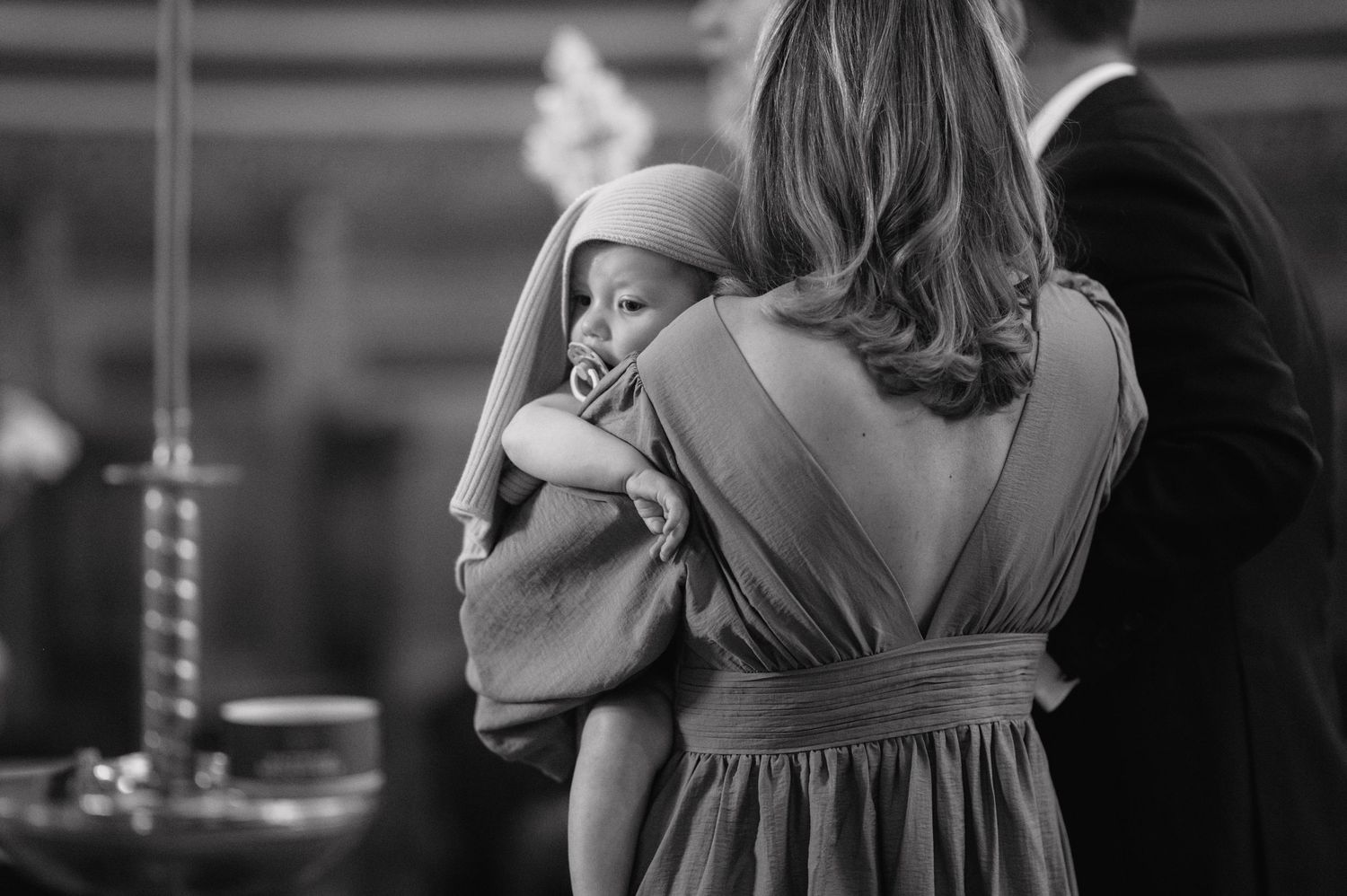 A black and white candid photo shows someone in a backless dress holding an infant during a formal event.
