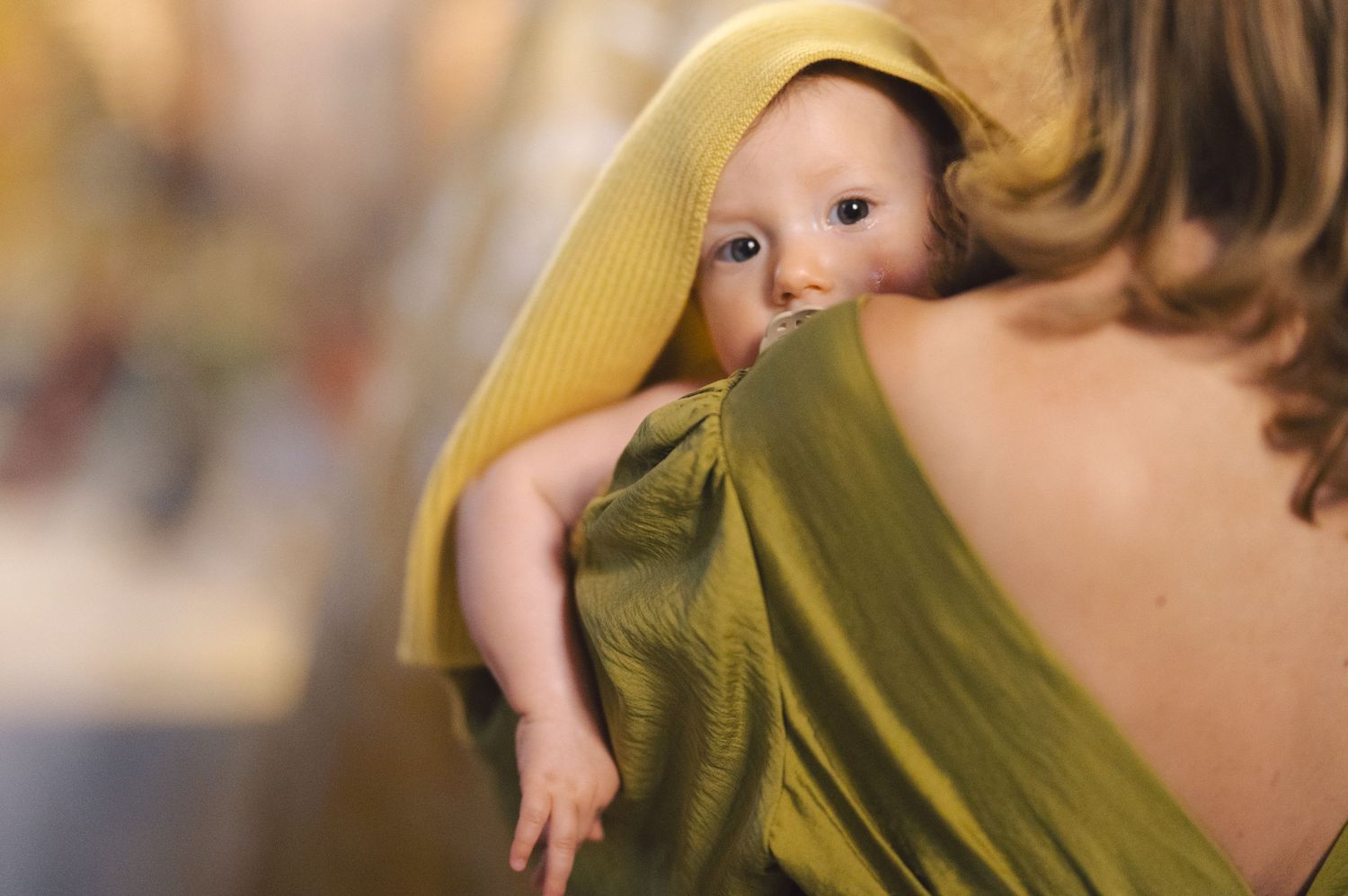 A baby peeks out curiously while being held in a yellow-green wrap against someone's back.