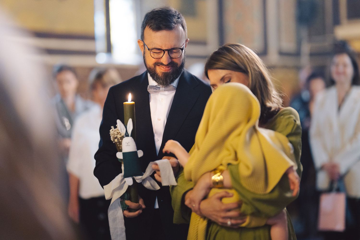 A church ceremony shows a person in glasses and bowtie performing a religious service while holding a child in yellow clothing.