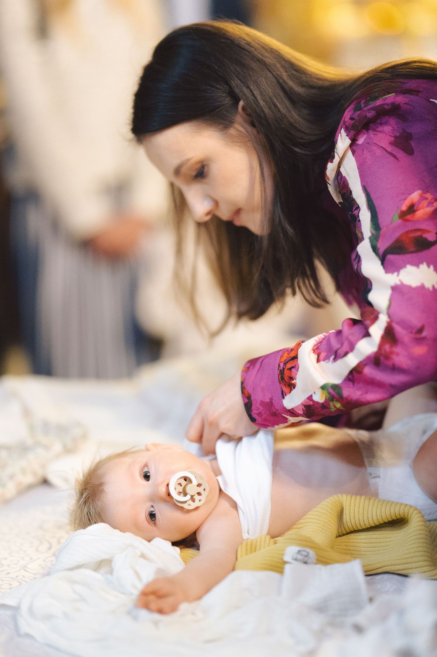 A mother in a purple floral top tends to a smiling baby wearing a pacifier laying on white bedding.