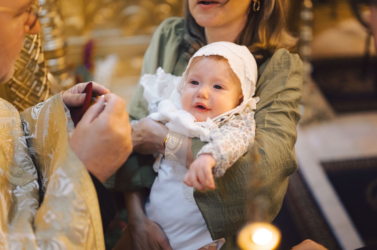 Baby in white christening gown being held during traditional baptism ceremony with warm candlelit ambiance.