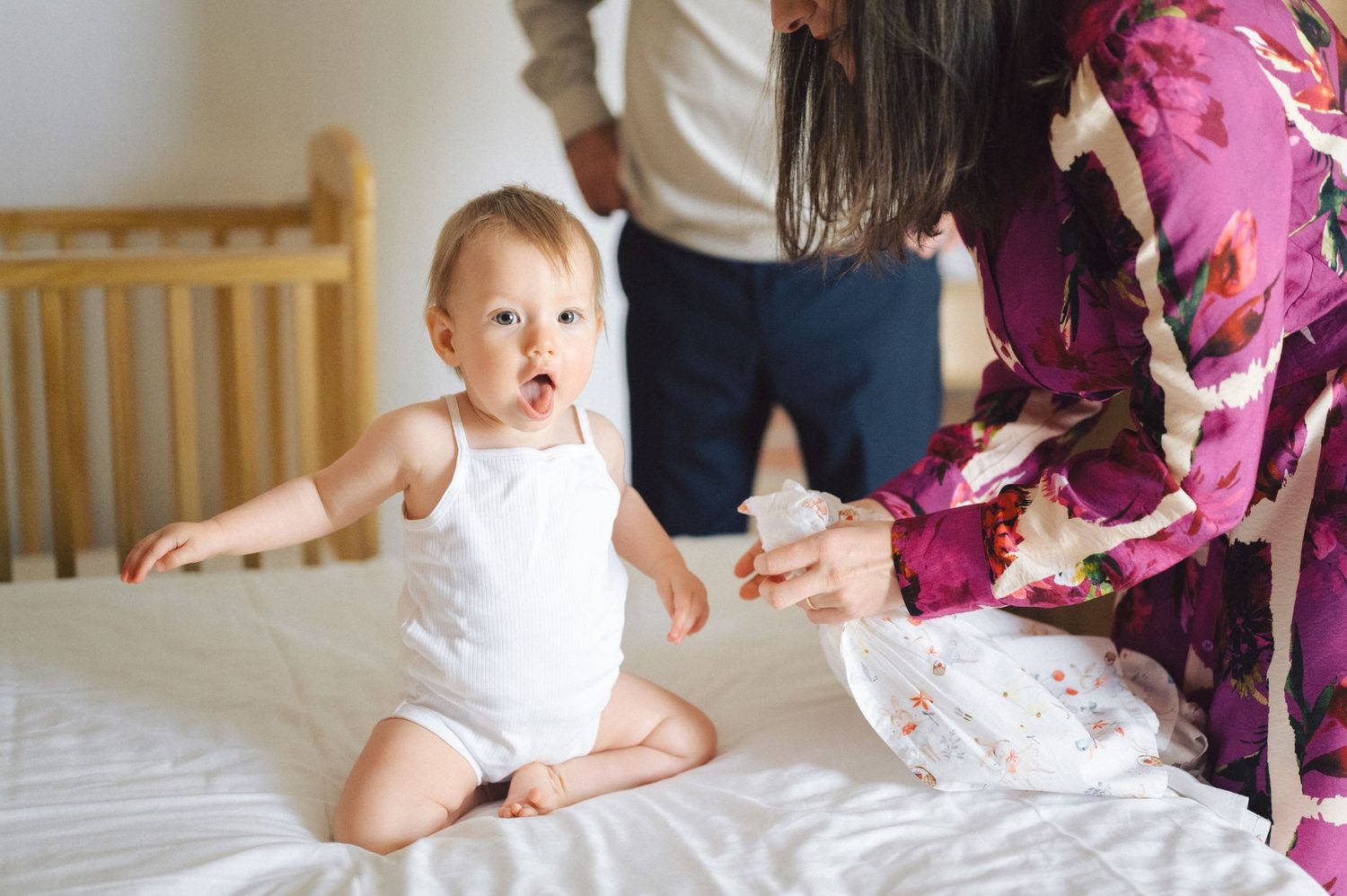 Young child in white attire sitting on bed with caretakers visible in casual and floral clothing nearby.