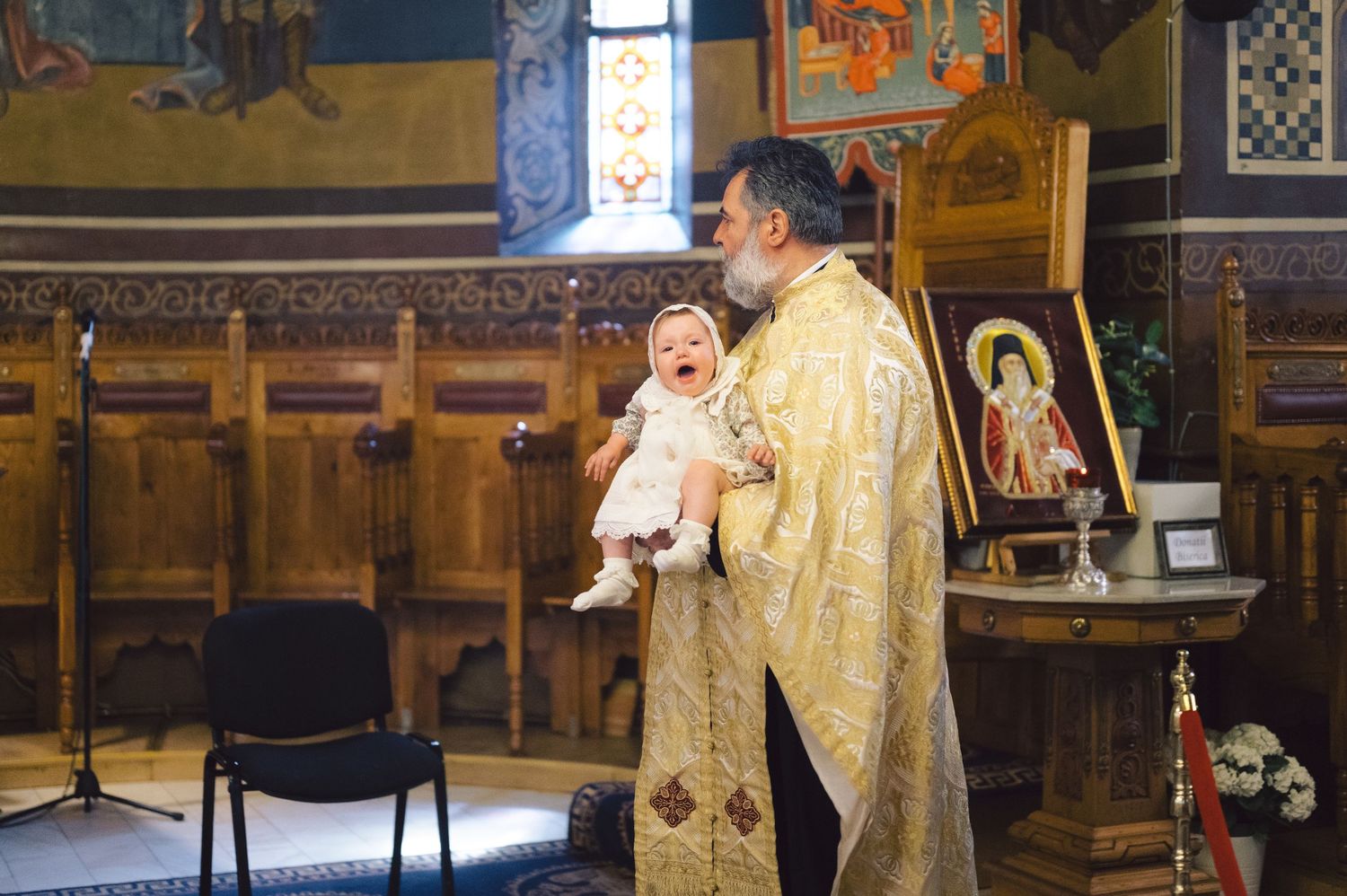An Orthodox priest in golden vestments performs a religious ceremony inside a traditional church with wooden panels and icons.