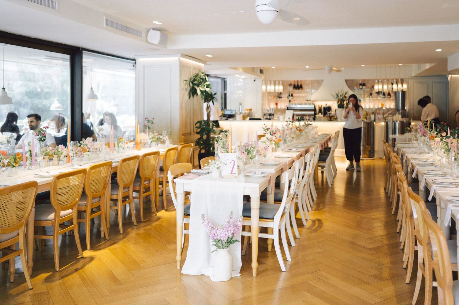 Elegant reception venue with long white tables, wooden chairs, and natural light streaming through large windows.
