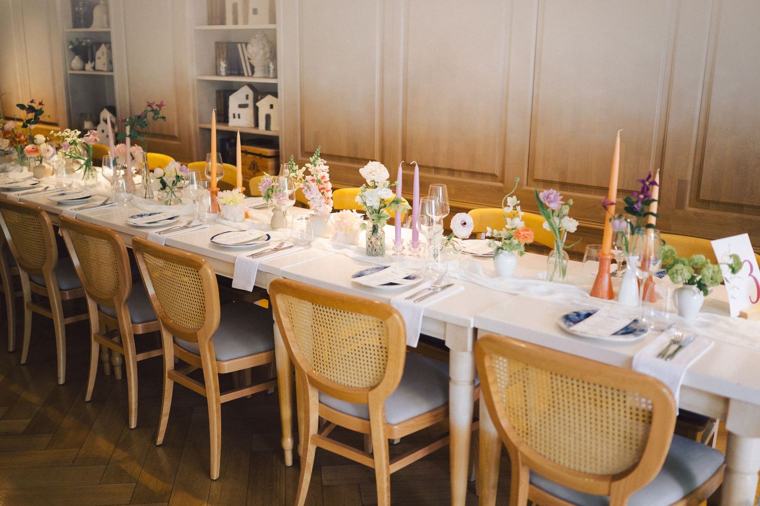 An elegant dinner table set with glowing candles, white linens, floral centerpieces and wooden chairs for a special occasion.