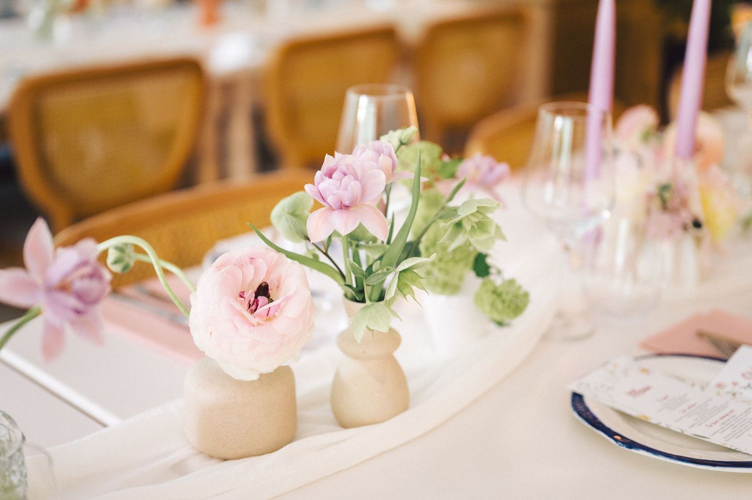 Small vases with delicate pink ranunculus and greenery decorate an elegant table setting with glassware.