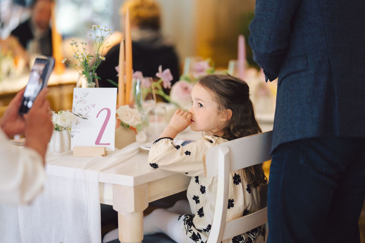 A young dinner guest in a cow print shirt sits at a decorated table while someone takes photos with a phone.