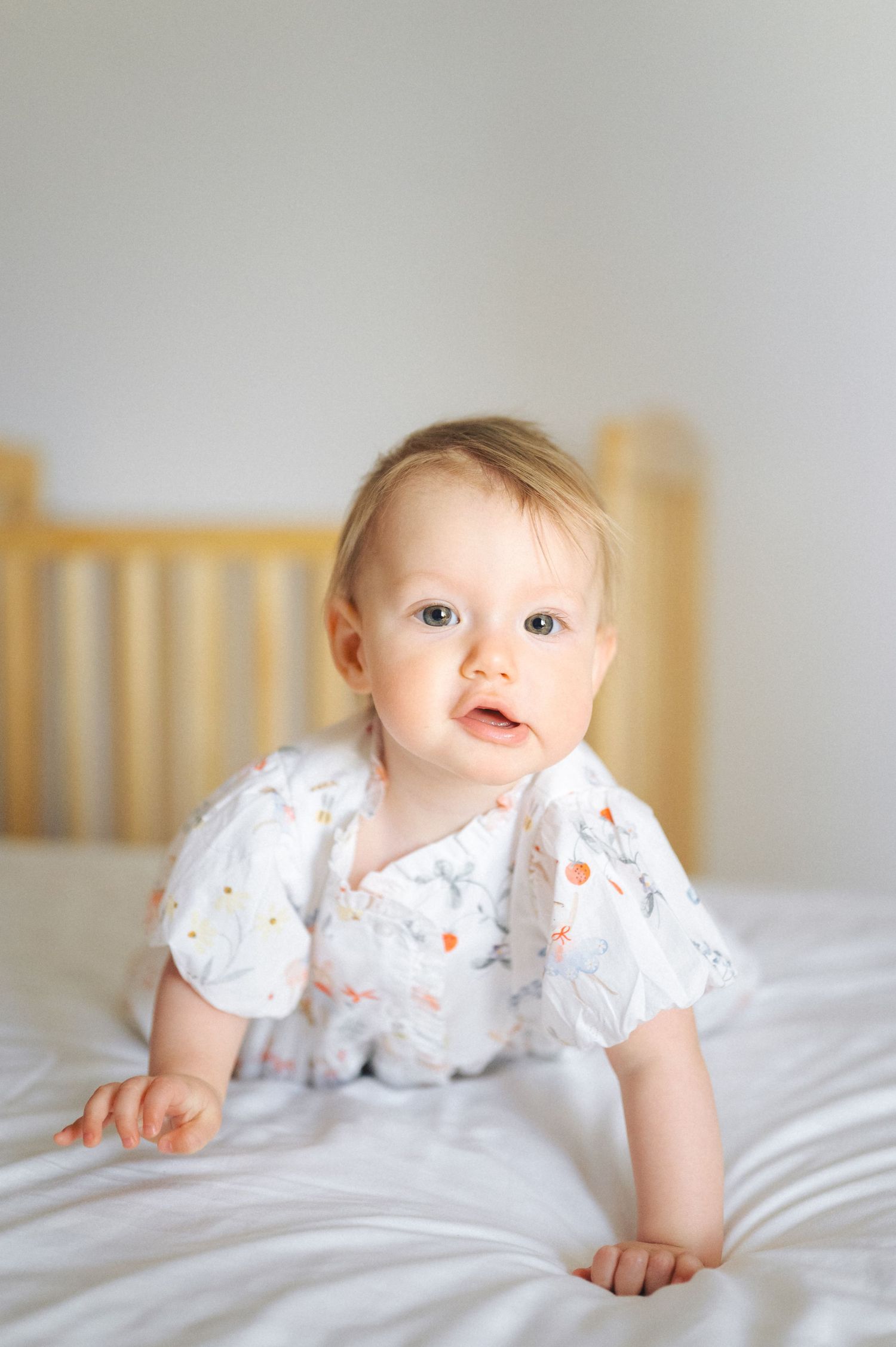 A baby in a white patterned onesie crawls on soft gray bedding with a wooden crib in the background.