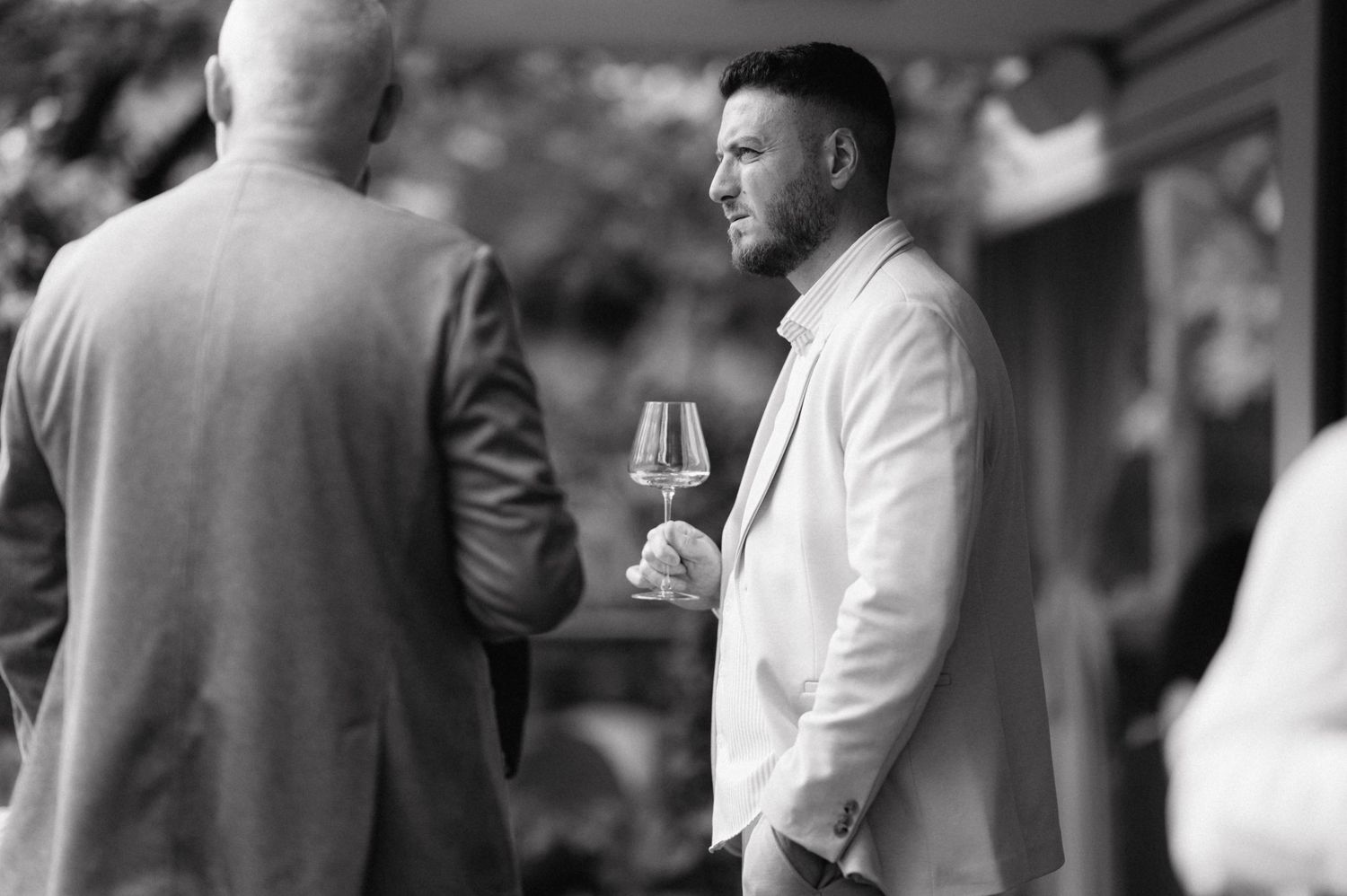 Two men in formal attire having a conversation while holding wine glasses in black and white photography.
