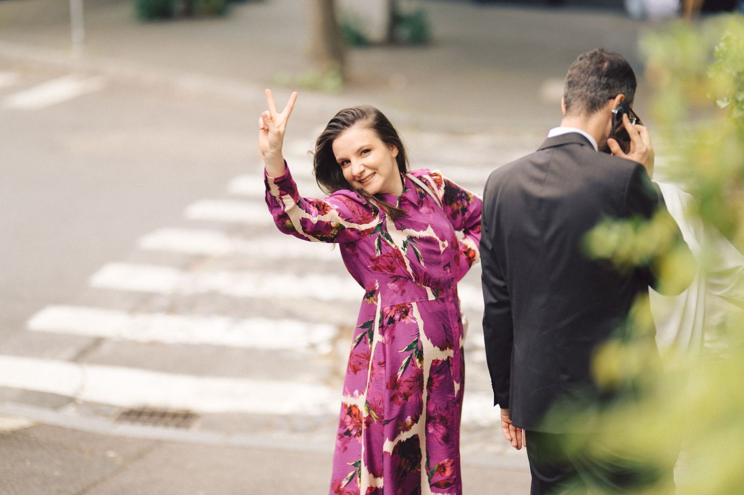 A person in a pink floral dress makes a peace sign while someone in a suit talks on the phone beside a crosswalk.