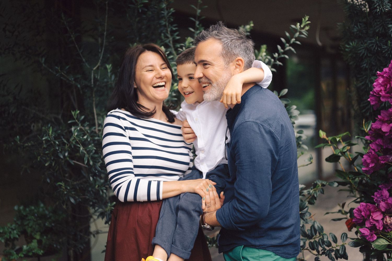 Candid portrait of three people laughing and embracing outdoors with pink flowers in the background.