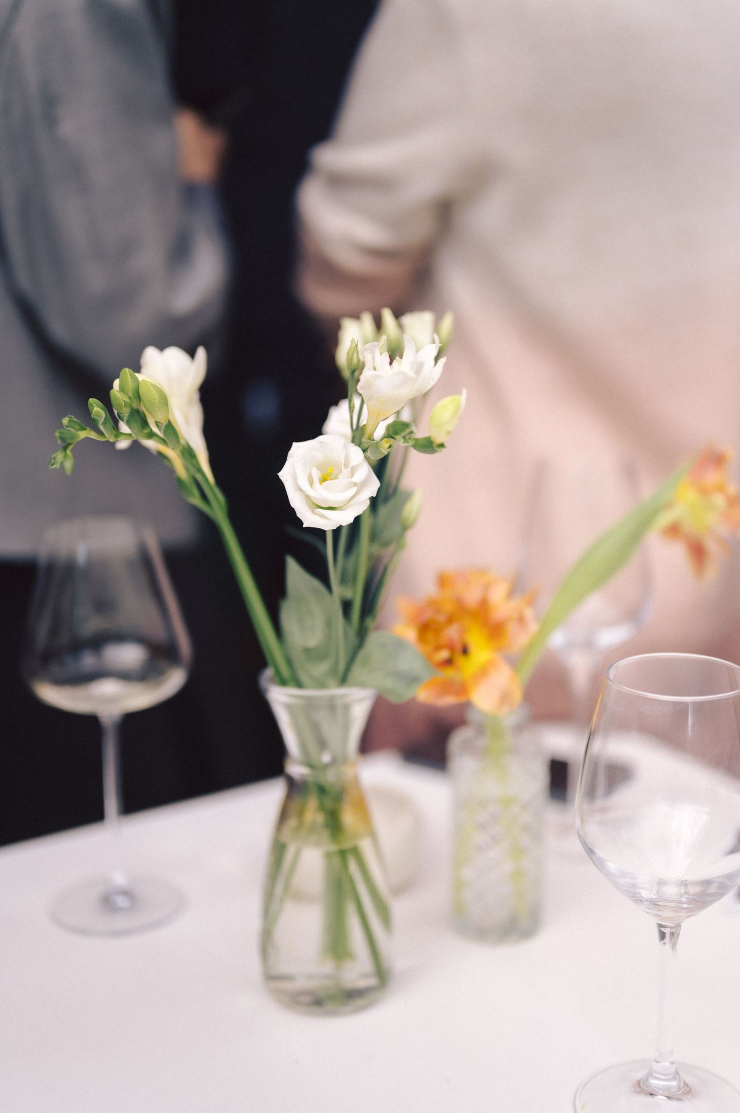 Simple white flowers and vibrant orange blooms in glass vases on white table with wine glasses, creating an elegant setting.