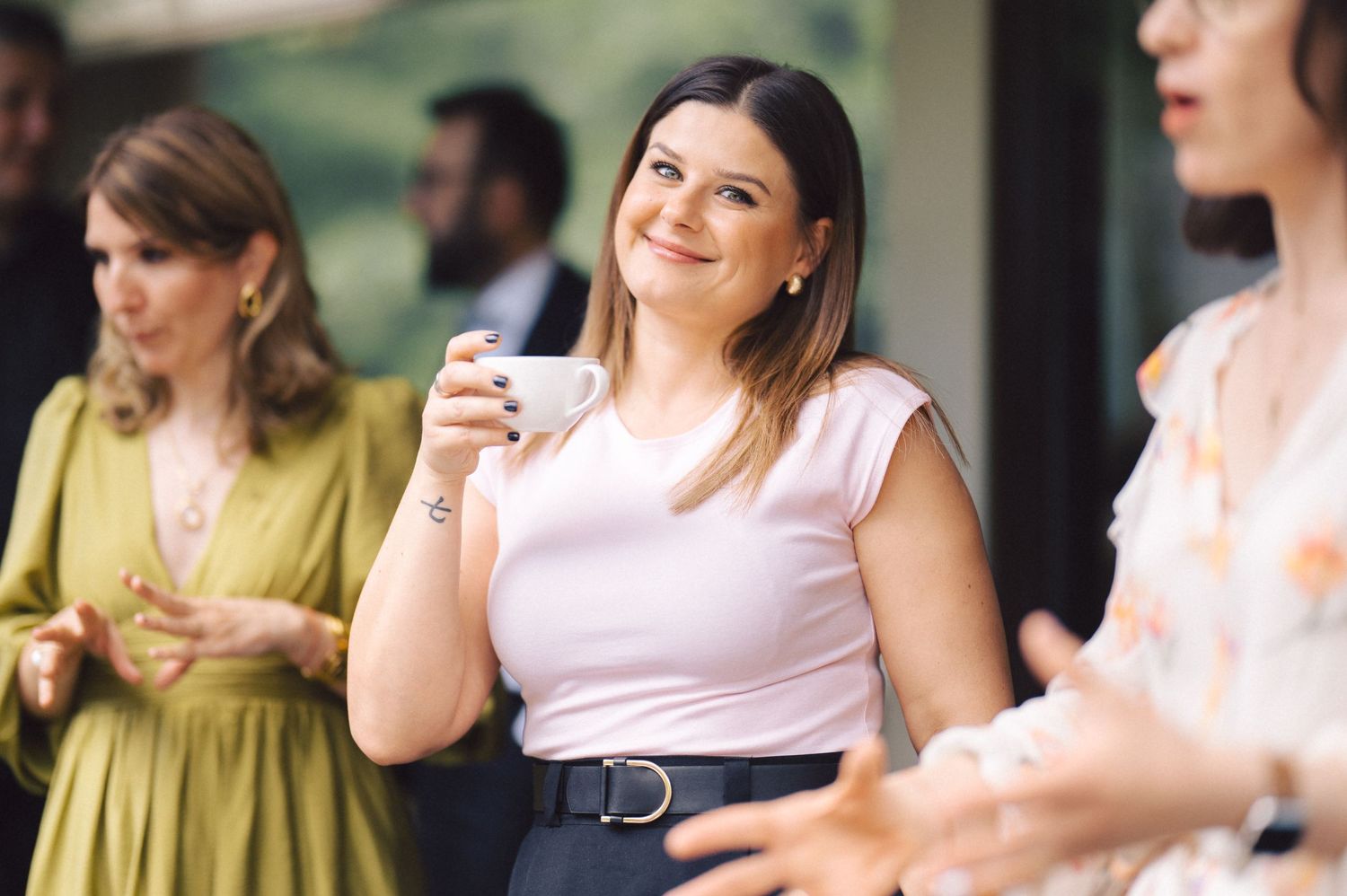 A person in a pink top and black belt holds a coffee cup while socializing at an outdoor gathering.
