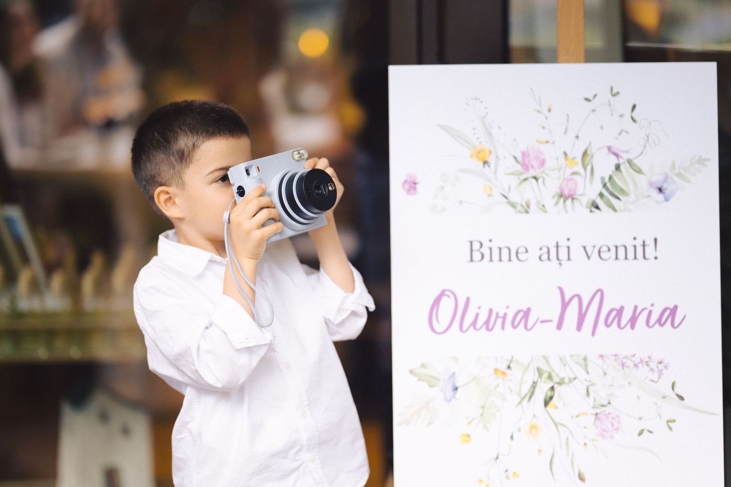 Young child in white shirt drinks from metal cup while standing near decorative invitation with pink text and floral design.