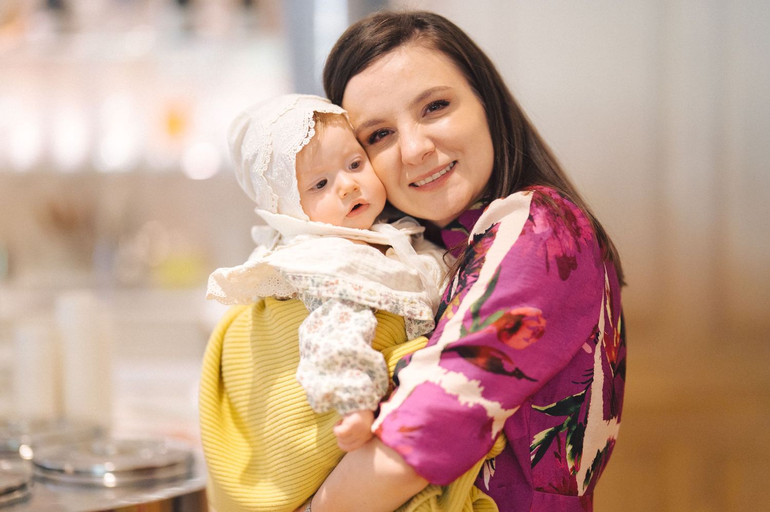 A parent dressed in a floral pink shirt holds a baby wearing a yellow outfit indoors.