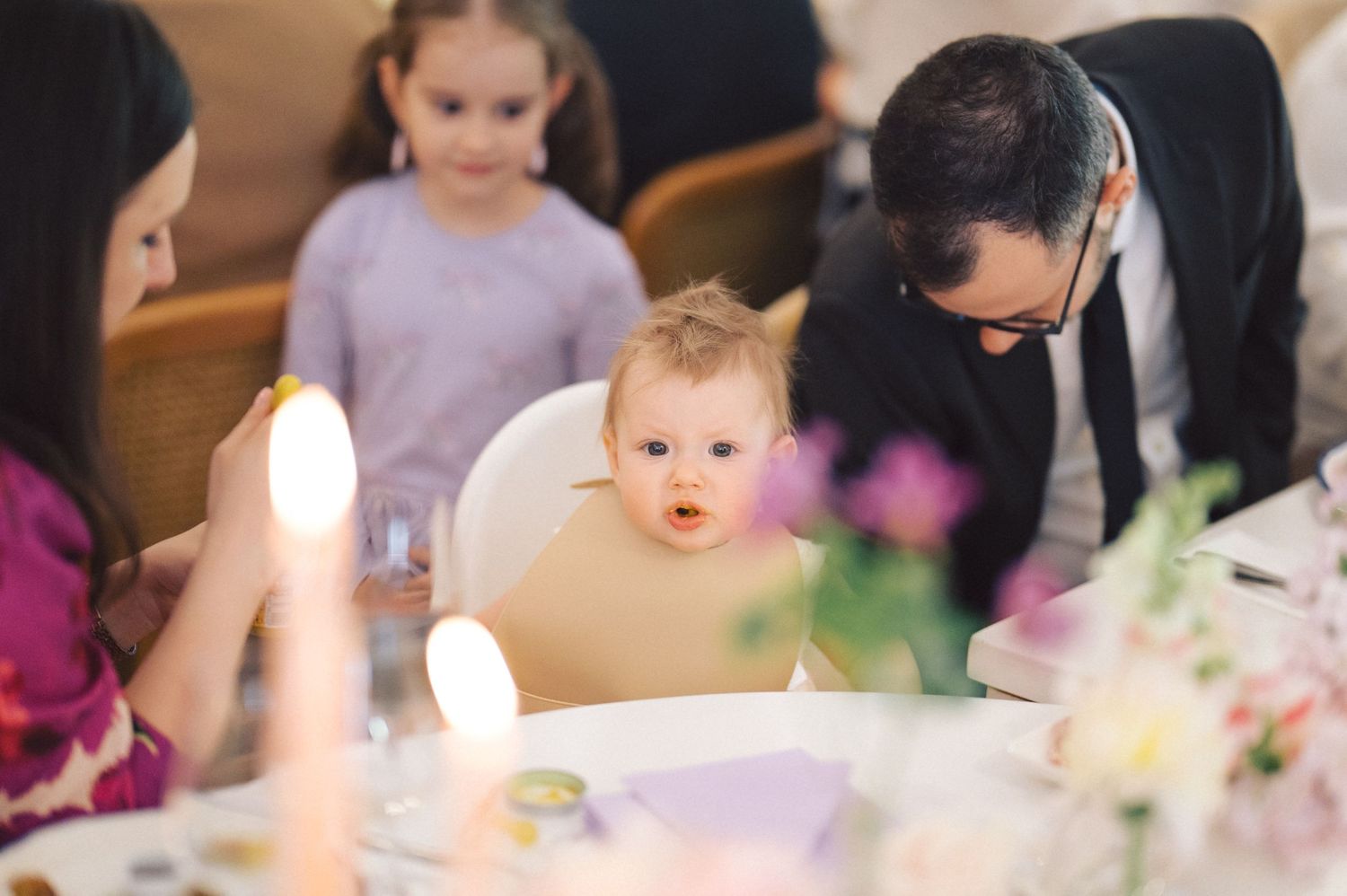 A small child in a high chair gazes in wonder at lit candles on a table while adults gather around during a celebration.