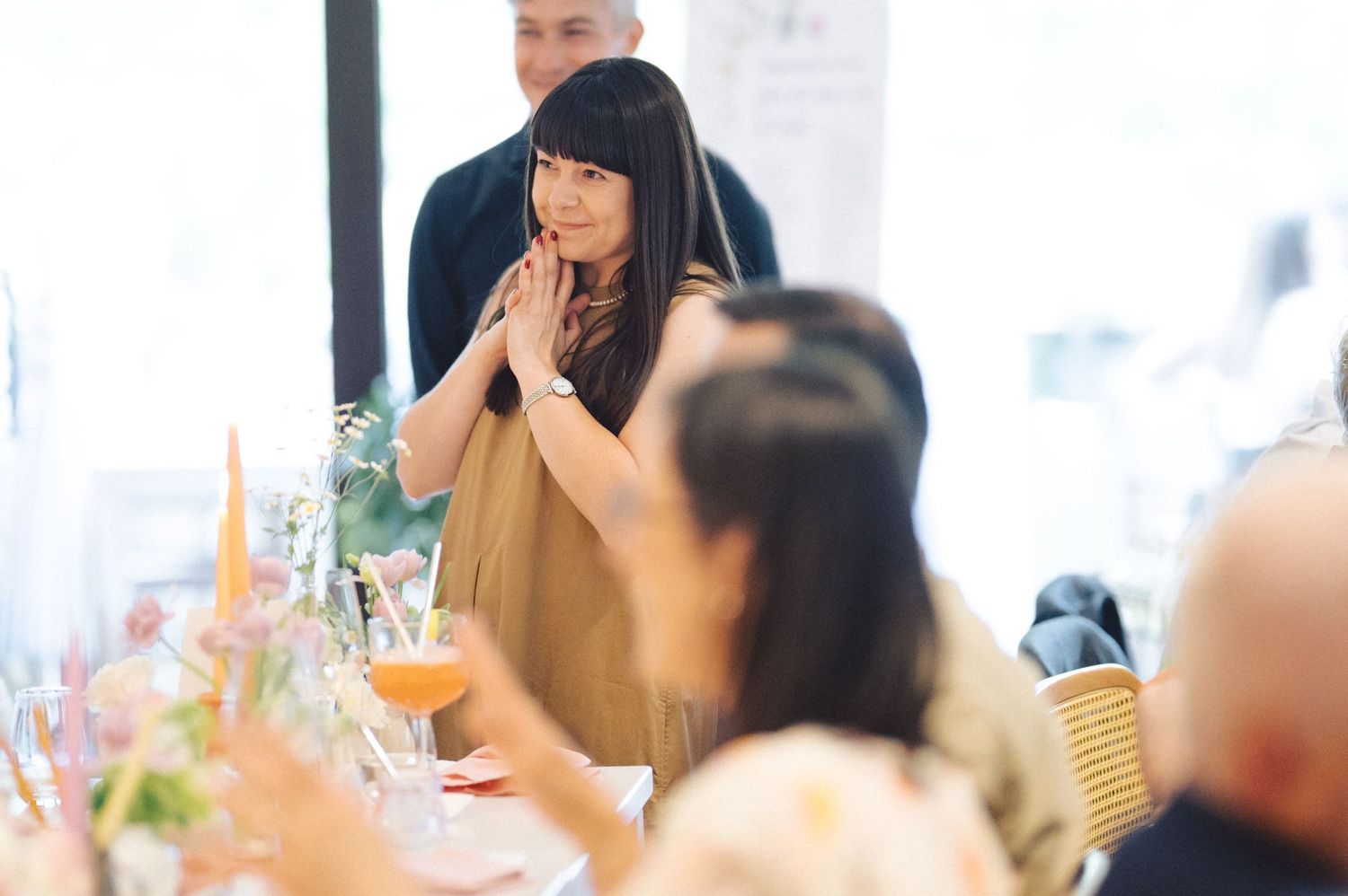 Person in a tan top sits attentively at a dining table during a social gathering.