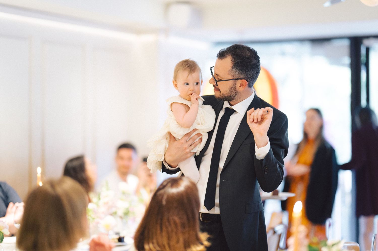 A speaker in a suit holds a baby while giving a speech at an elegant indoor event with guests seated at tables.