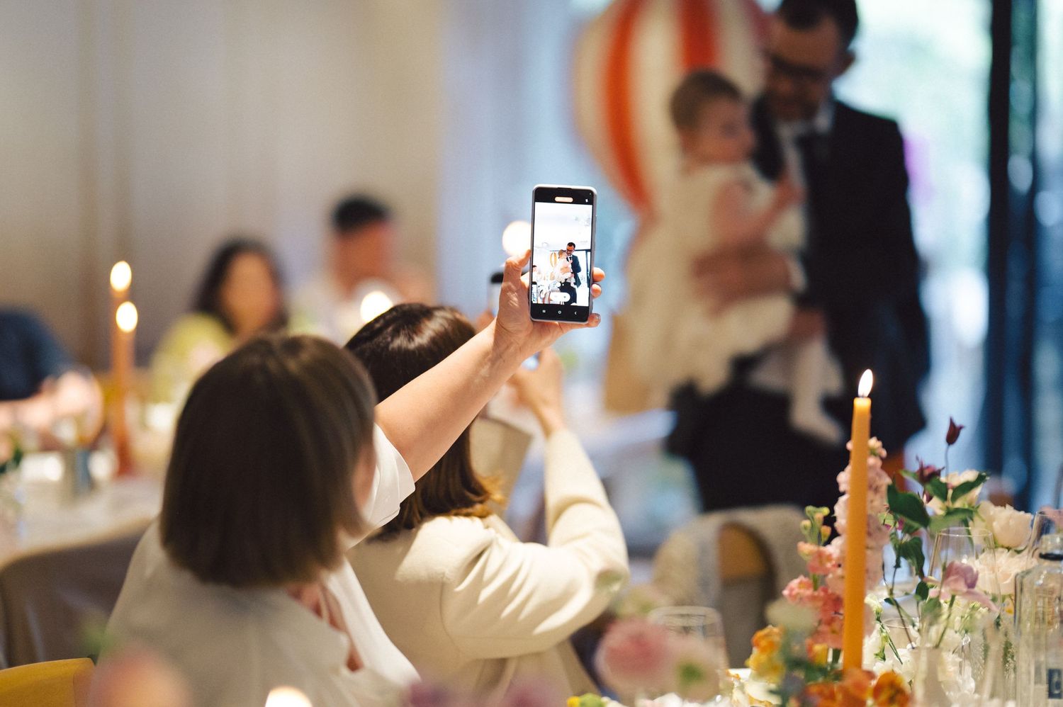 A guest holds up a smartphone to take photos during a celebratory dinner event with a candle lit table setting.
