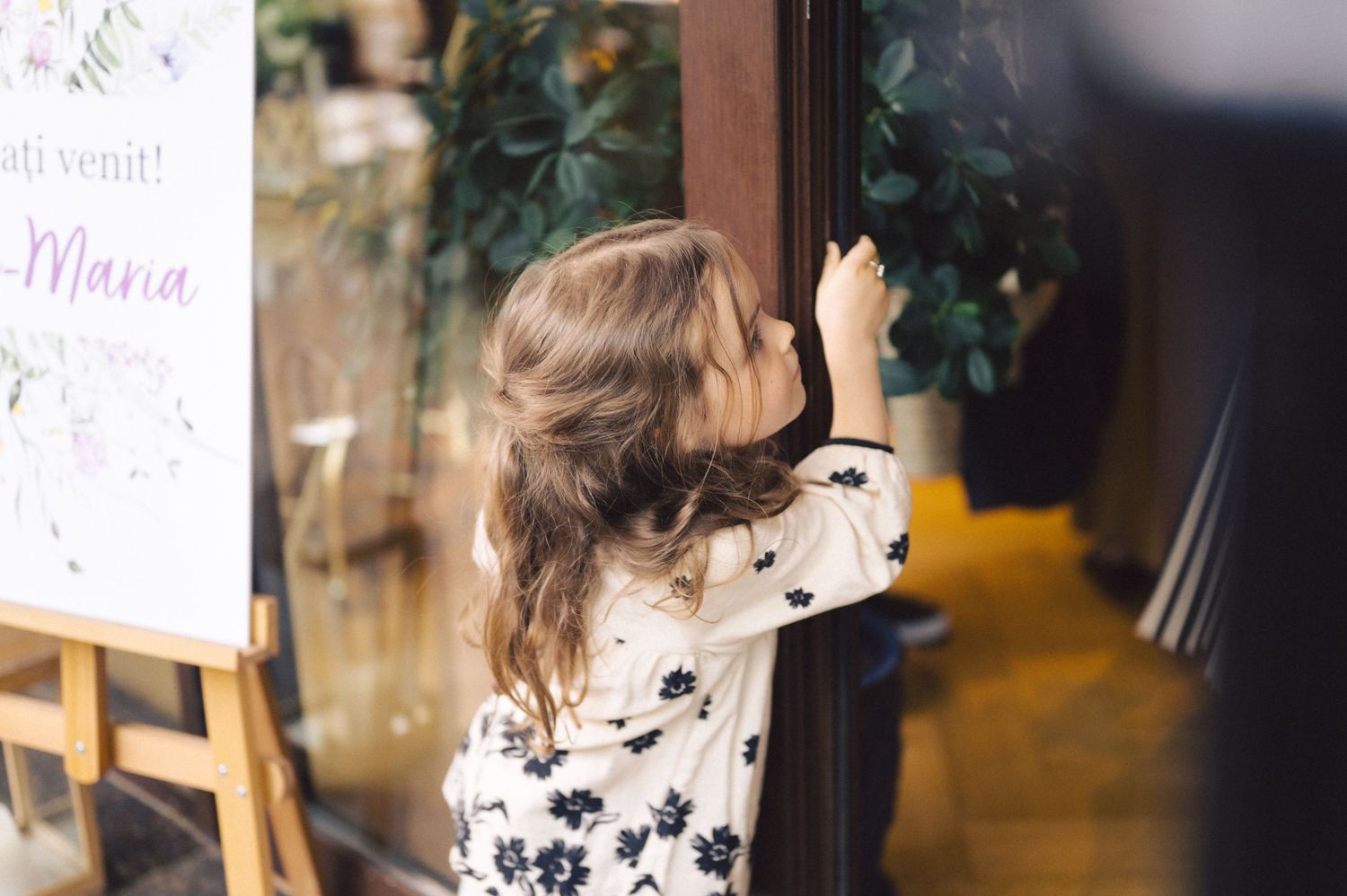 A child in a floral dress stands on tiptoes peering through a window into a shop.