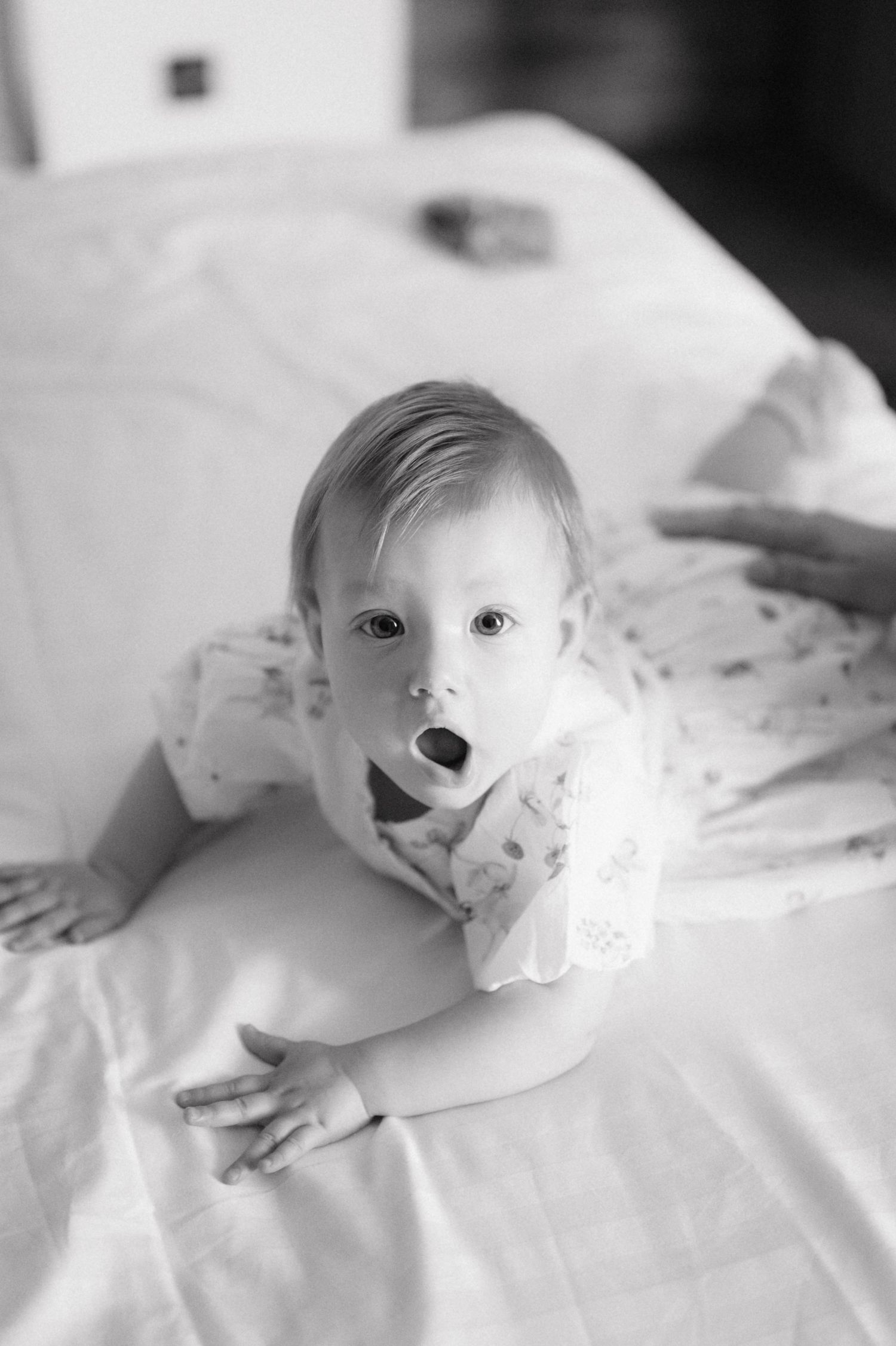 A black and white portrait of a small baby with wide eyes lying on white bedding making a surprised expression.