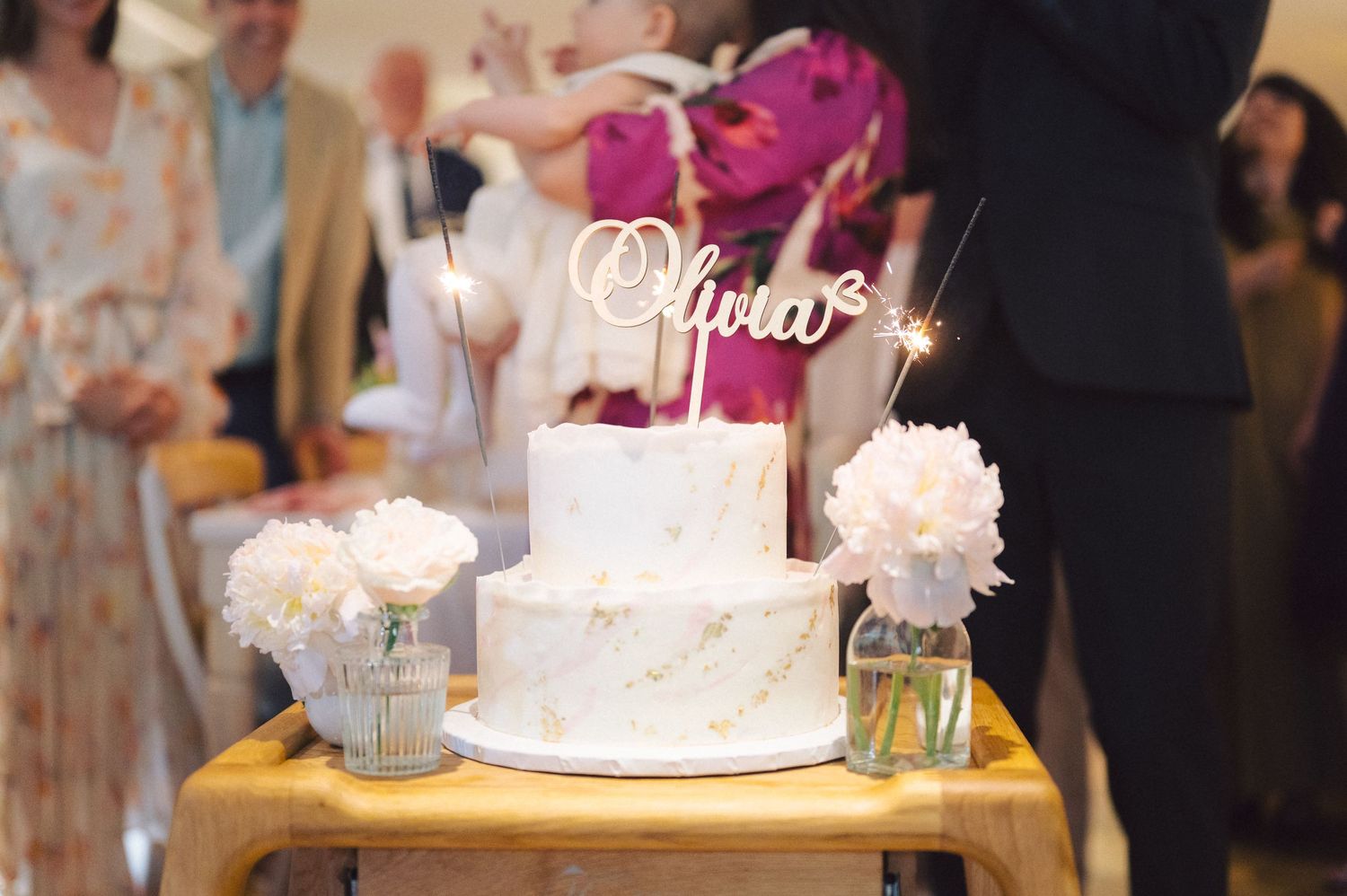 Elegant white tiered wedding cake with gold signage topper displayed on wooden table with floral decorations.