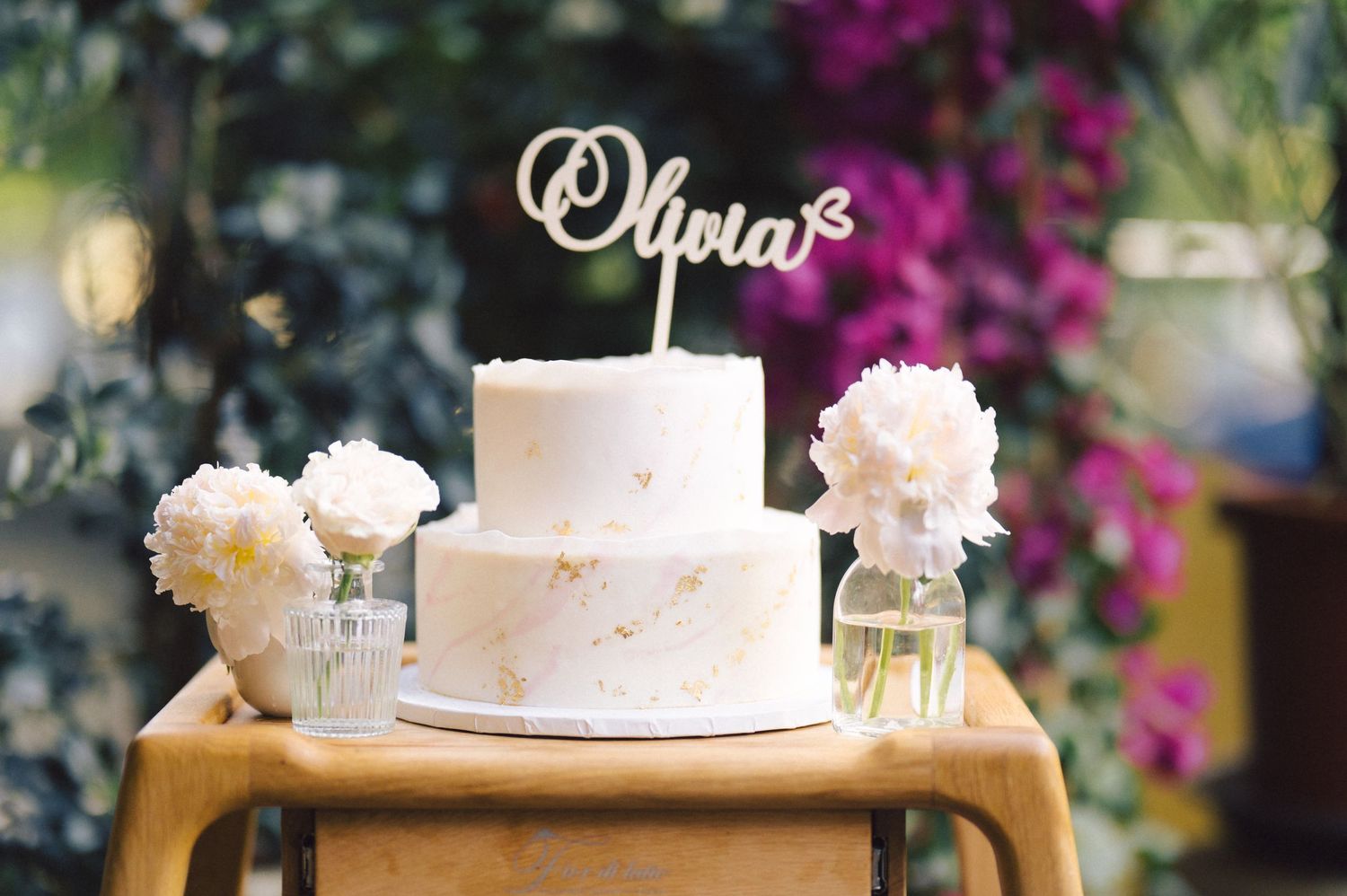 A white two-tier cake decorated with fresh flowers and a custom name topper sits on a wooden stand.