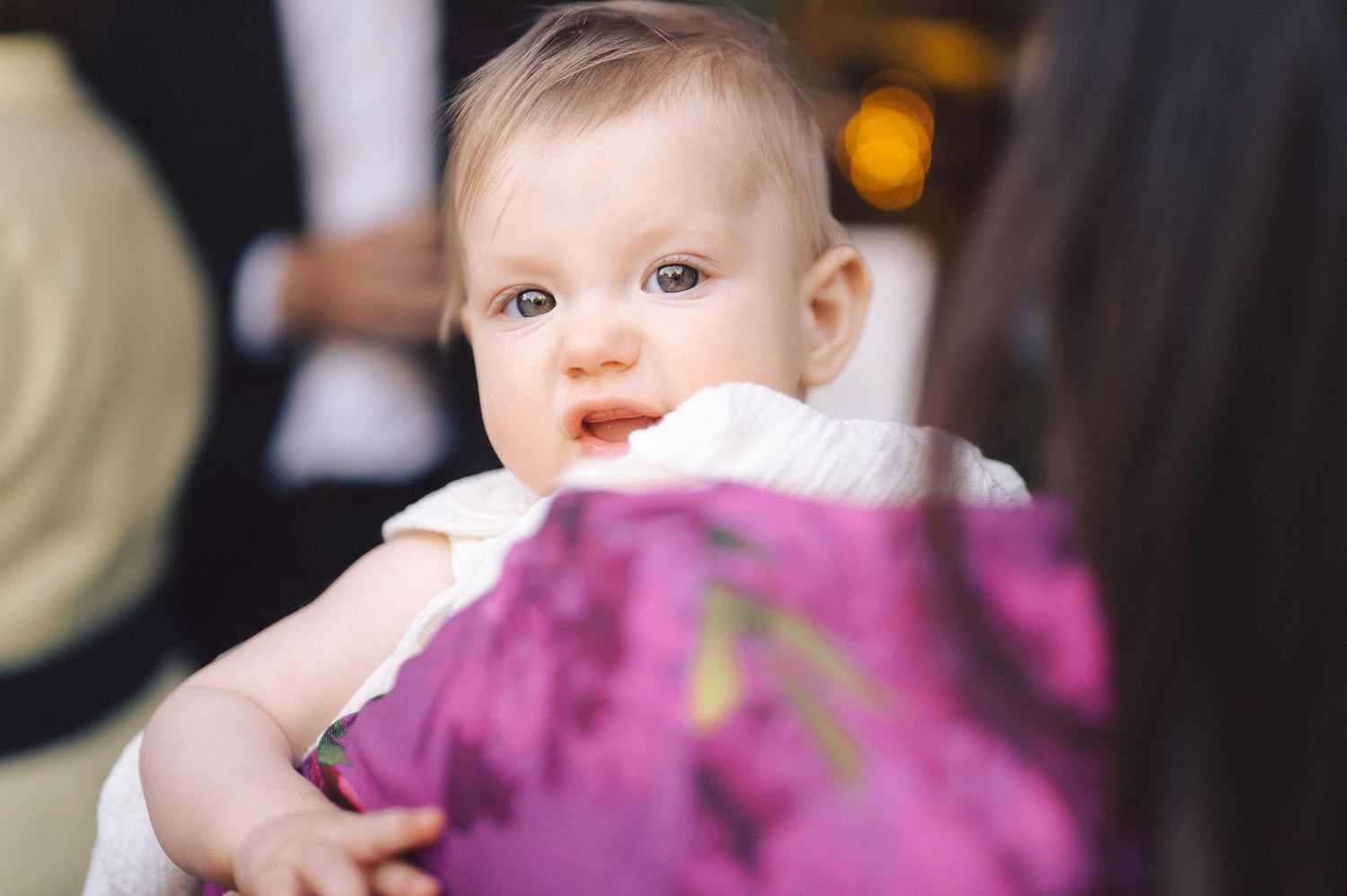 A baby wearing white attire at a celebratory event with purple decor in the background.