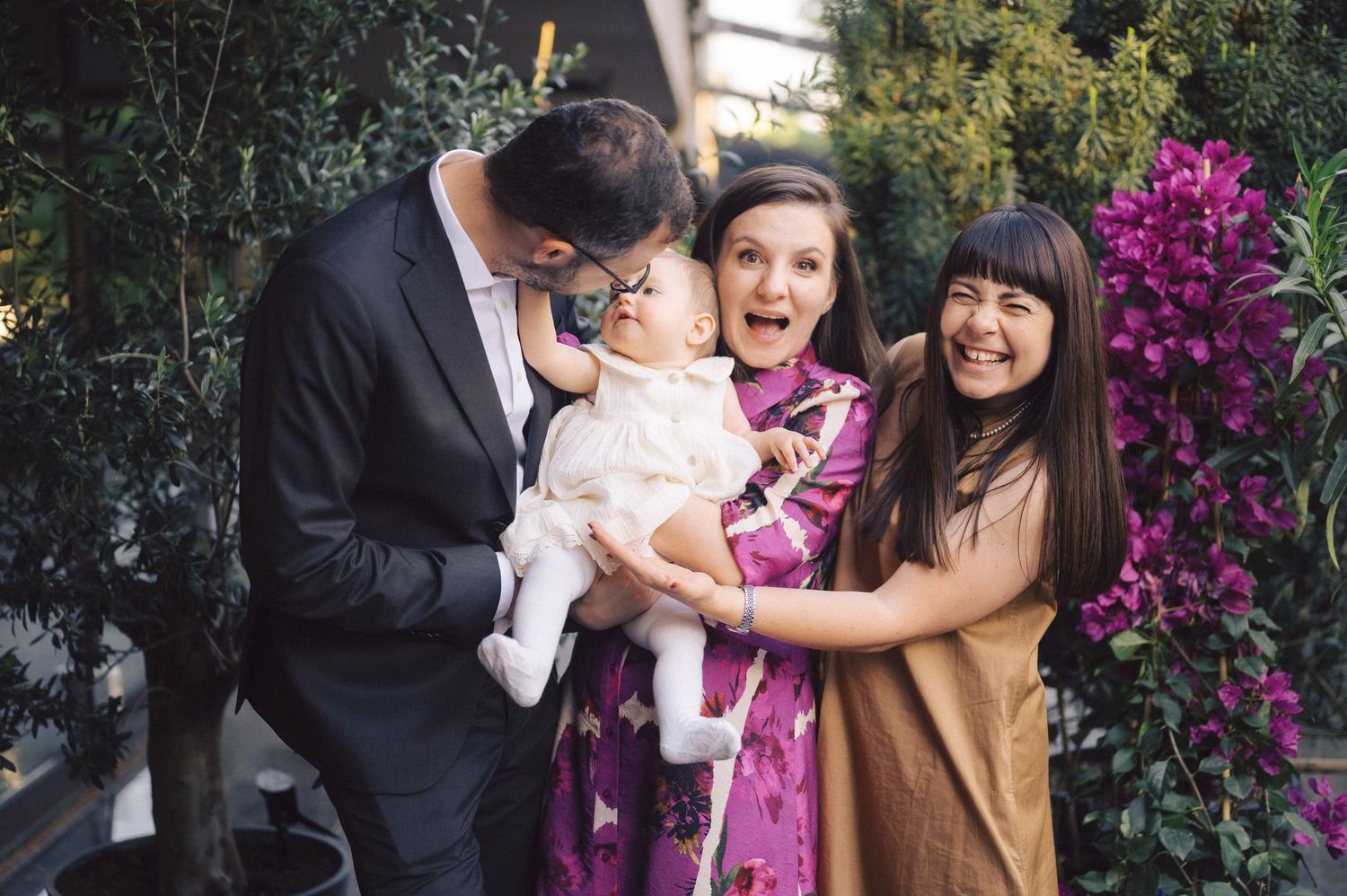 Three people share a joyful moment with a baby while standing in a garden setting with vibrant purple flowers.