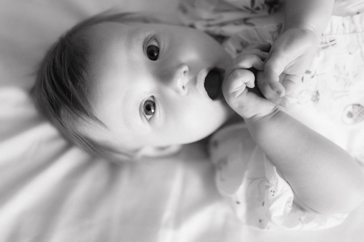 A little baby lies on white sheets looking up with wide curious eyes in black and white photography.