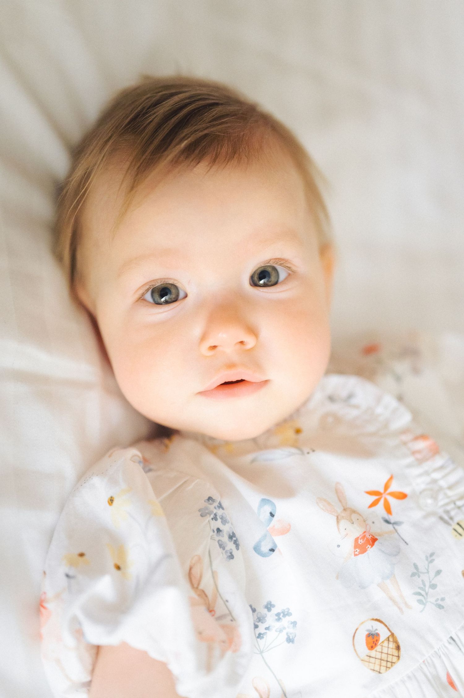 Baby wearing white patterned onesie lying on light colored bedding looking up with bright eyes and sweet expression.