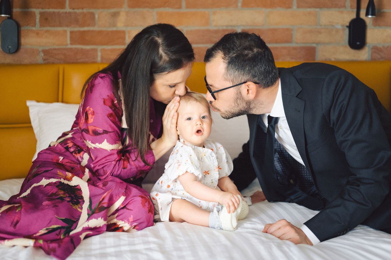 Family shares tender moment while relaxing on bed with young child in formal attire.