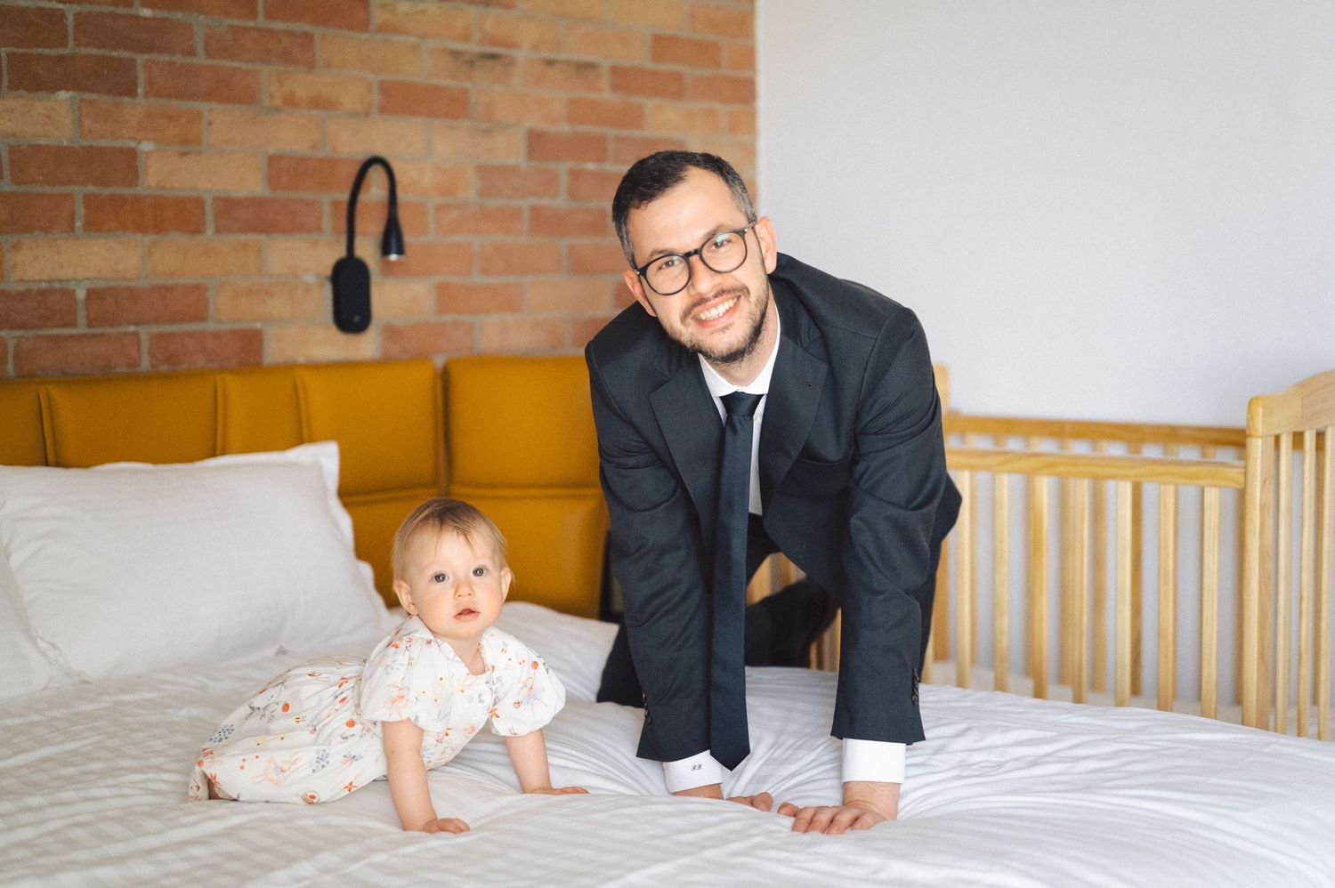 A small child in white clothing and adult on a bed with orange headboard and exposed brick wall background.