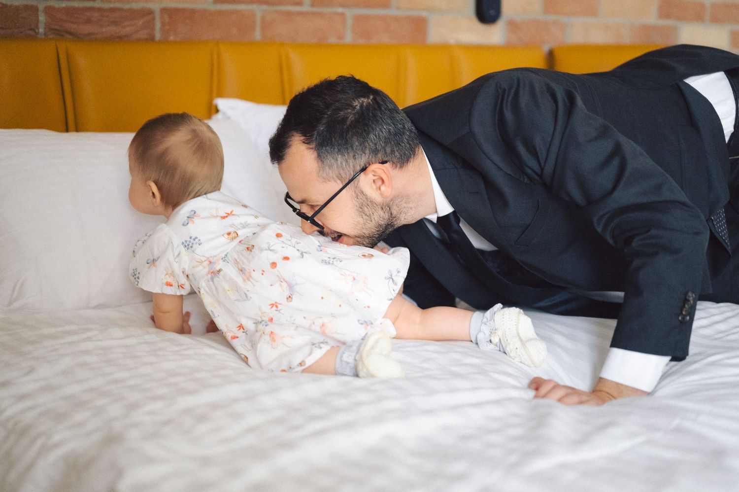 A baby in white pajamas crawls on a beige couch while someone in a black suit leans down playfully.