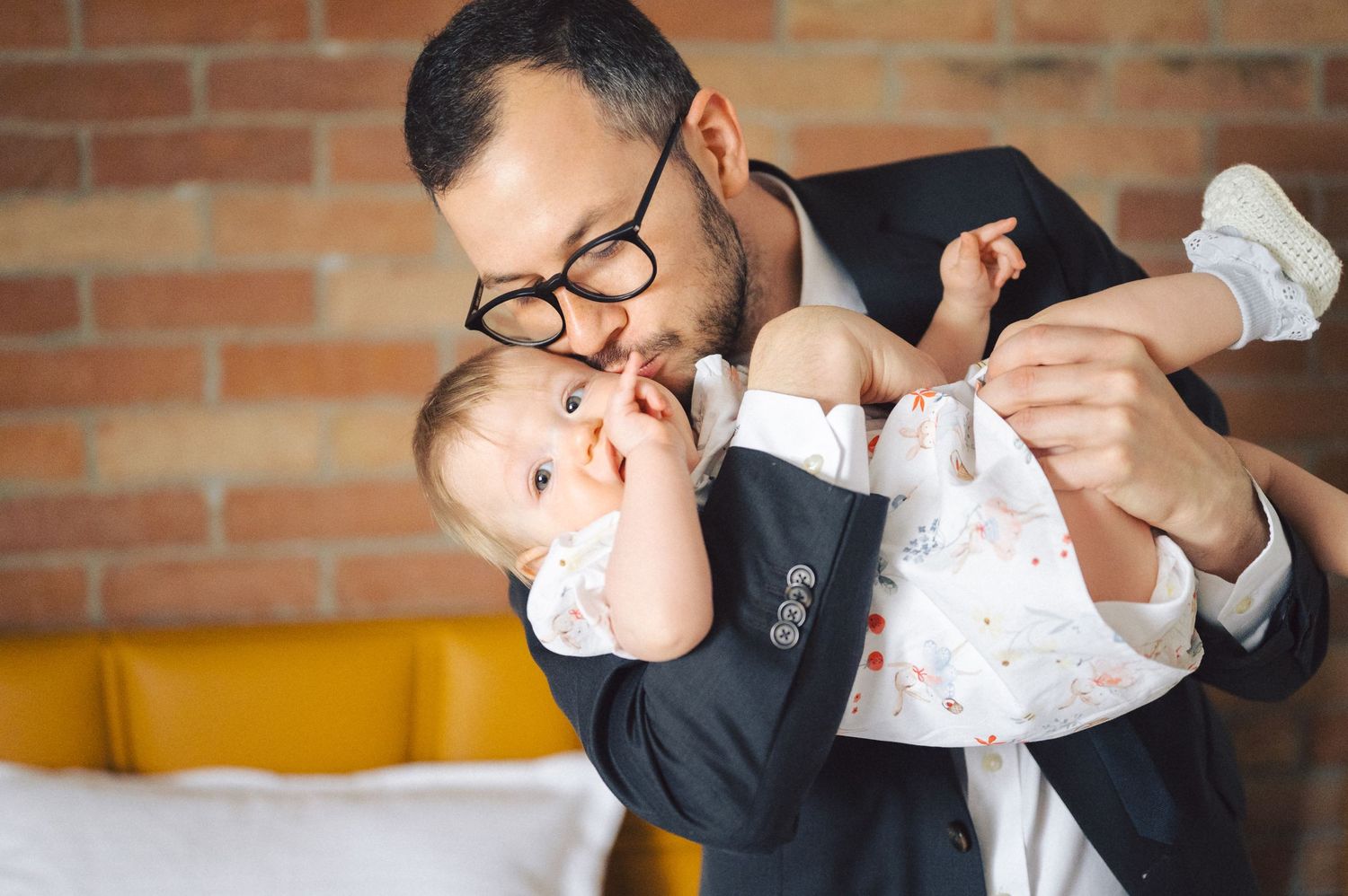 A playful moment between a father wearing glasses and suit jacket as he holds and entertains a smiling baby in white clothes.
