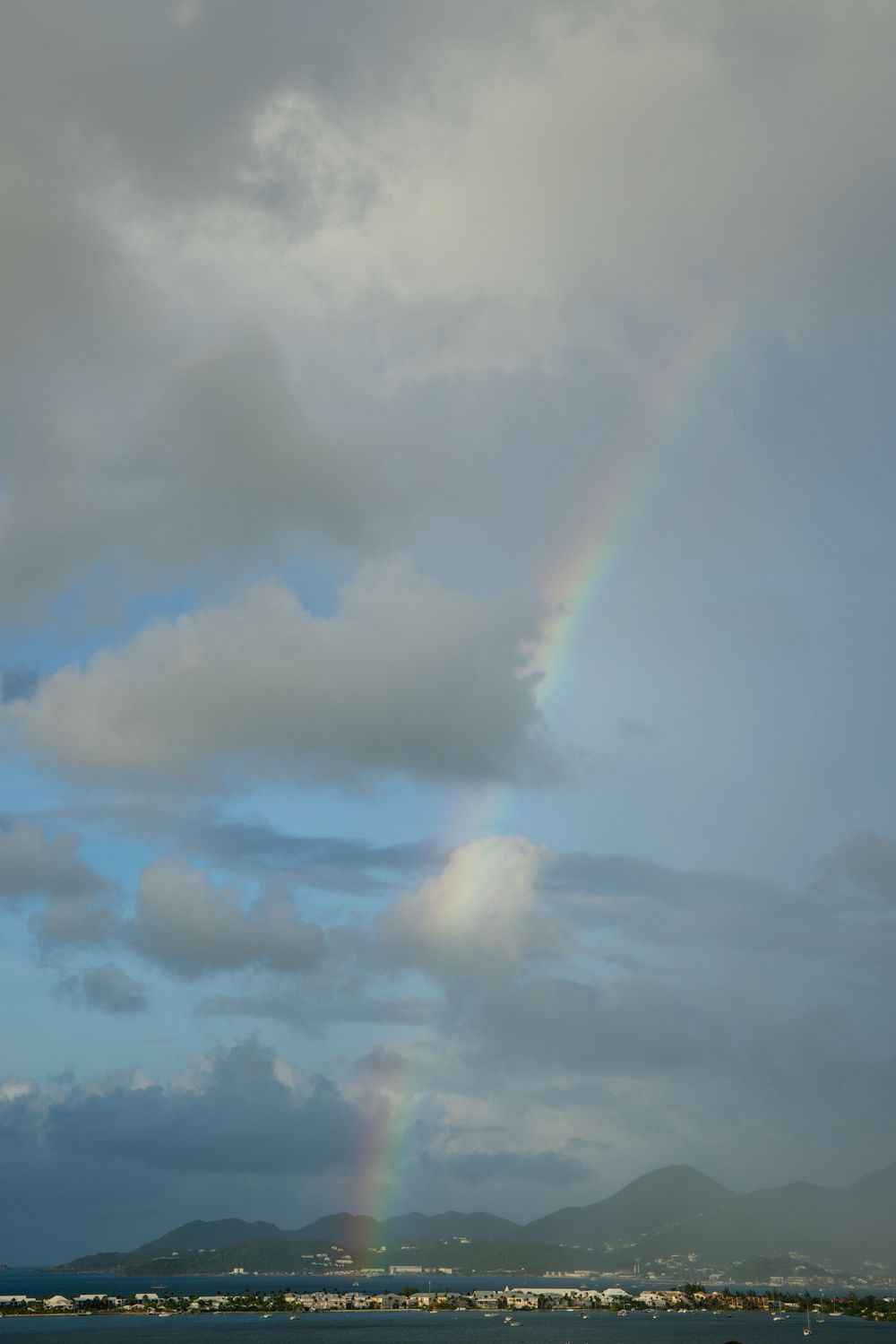 A vibrant rainbow arches over mountainous Caribbean coastline with cloudy skies and coastal buildings below. Saint Maarten