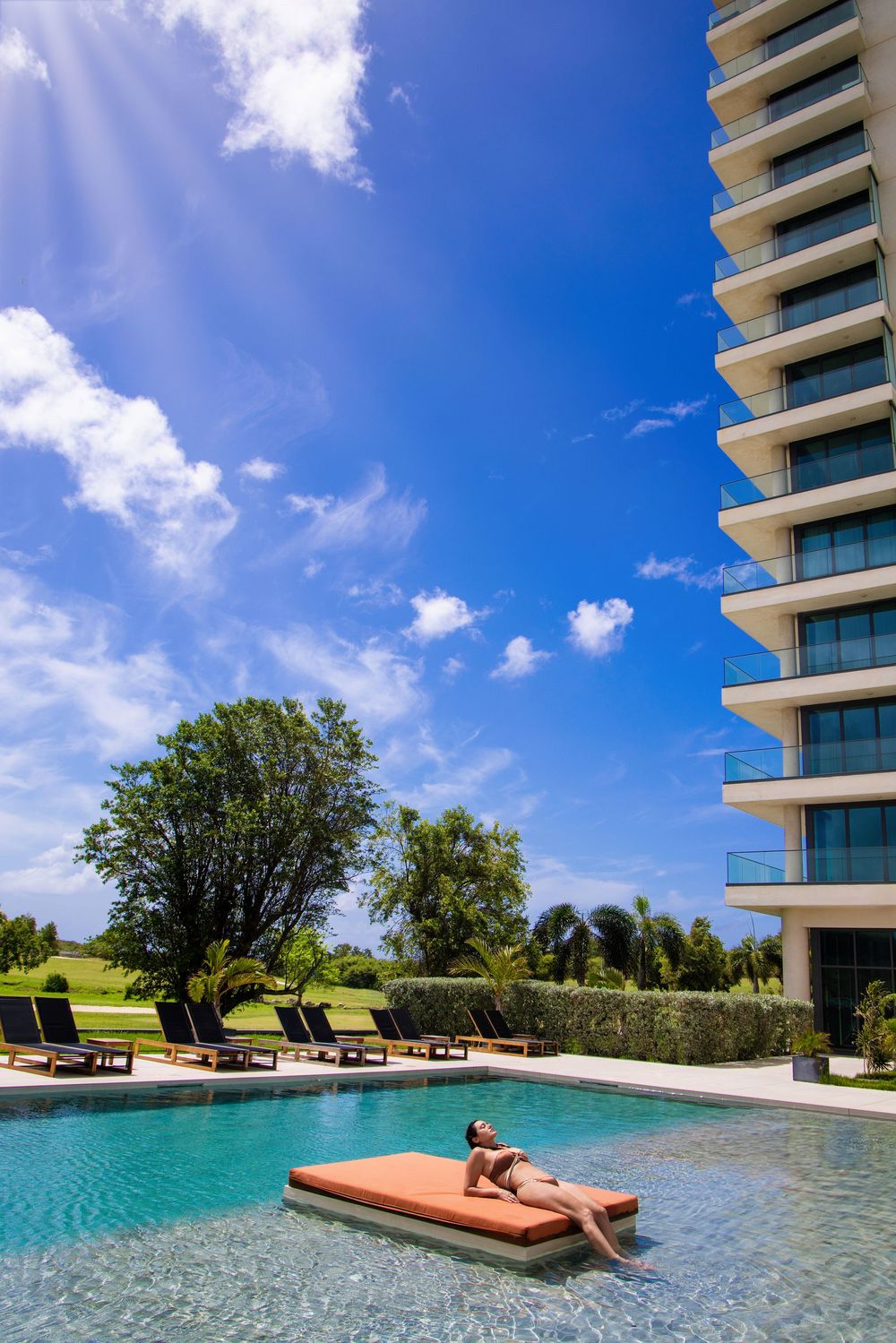 Person relaxing on floating lounge in luxury hotel pool with high-rise building and blue sky backdrop. Fourteen Mullet Bay Saint Maarten