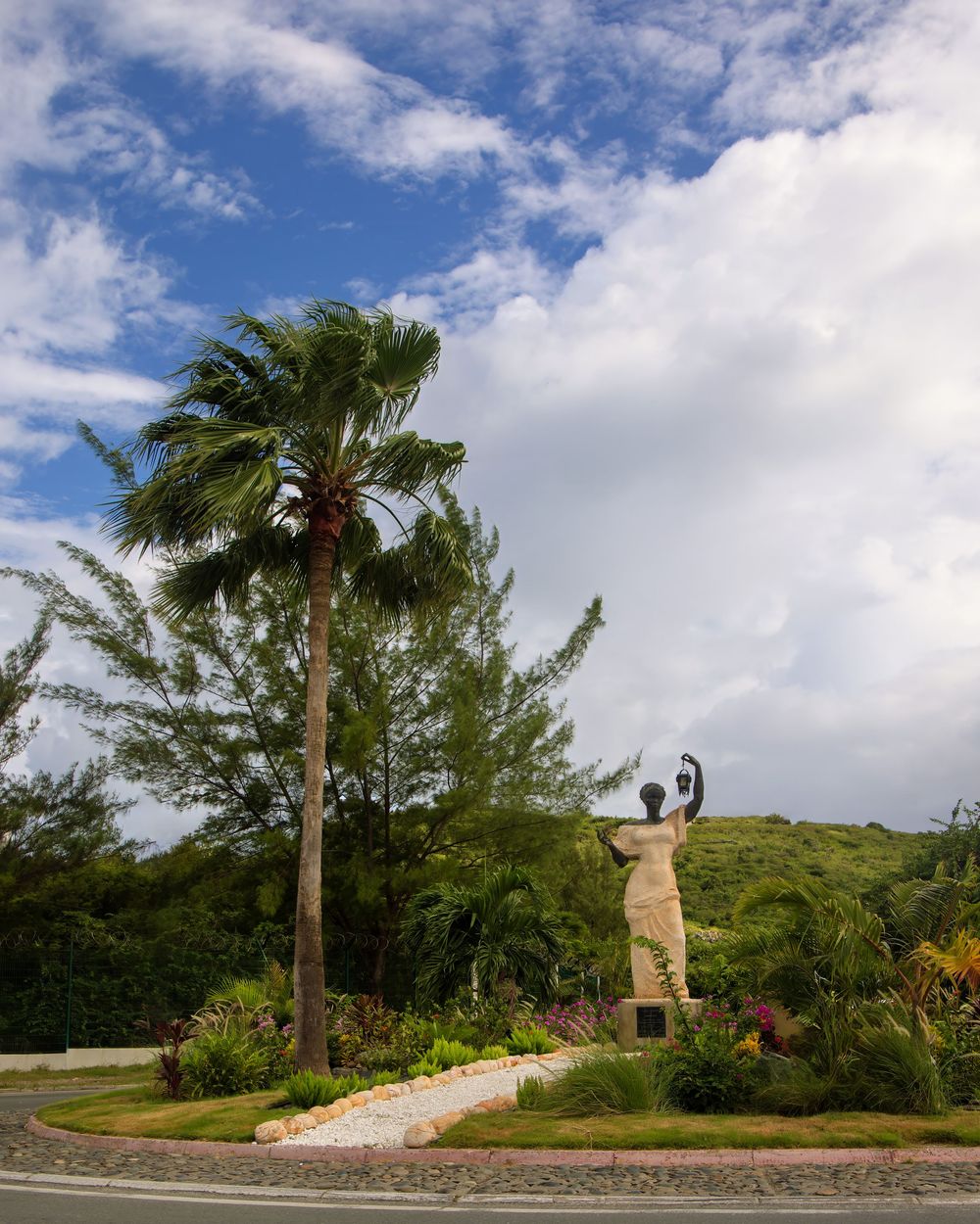 A tall palm tree stands beside a statue on a hill against a blue sky with white clouds. Saint Maarten
