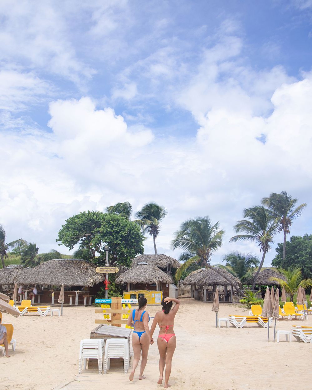 Two beachgoers enjoy a tropical resort setting with thatched huts, palm trees and yellow lounge chairs on a sandy beach. Saint Maarten