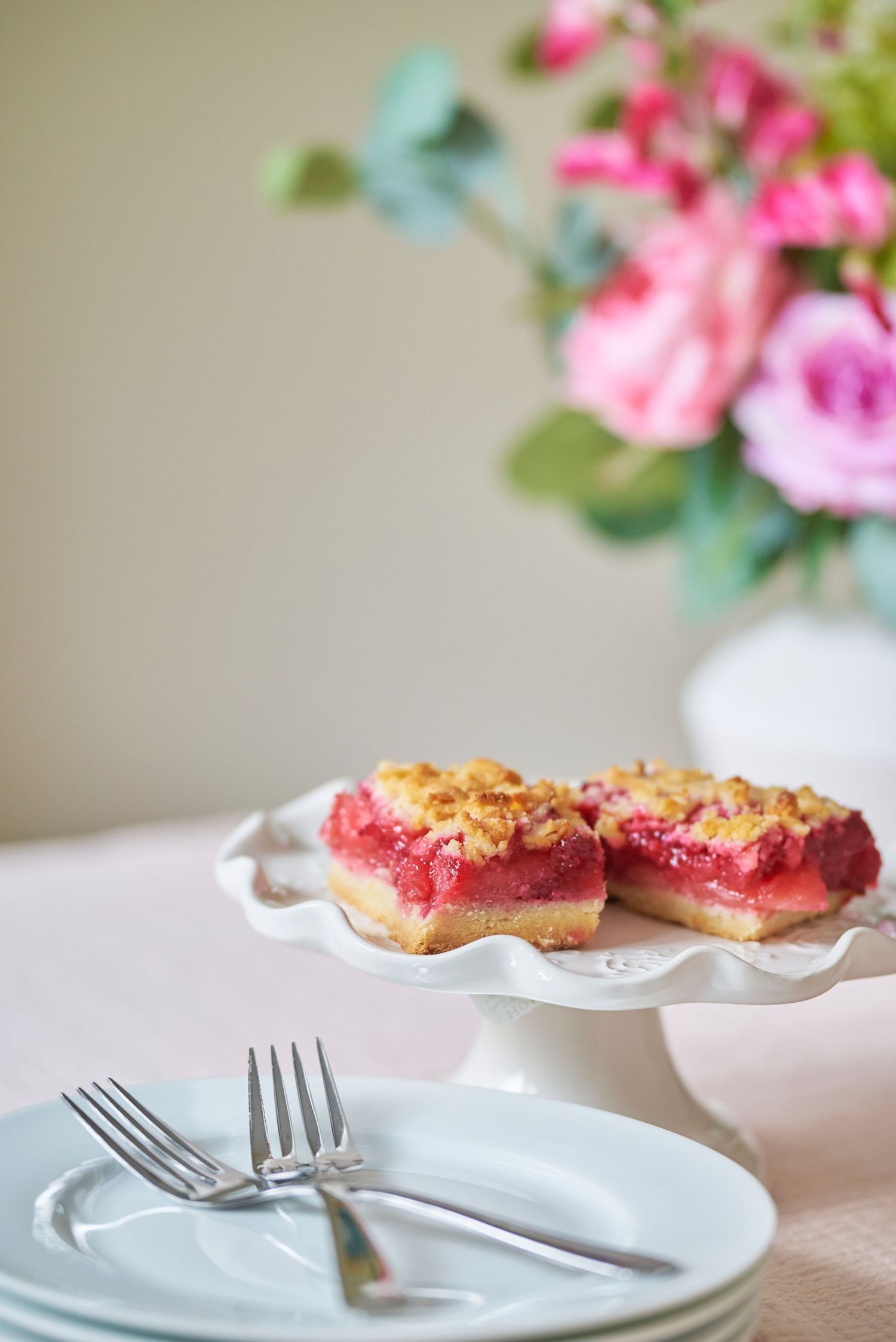 Pink dessert bars with crumb topping on a white scalloped cake stand next to forks and fresh flowers.