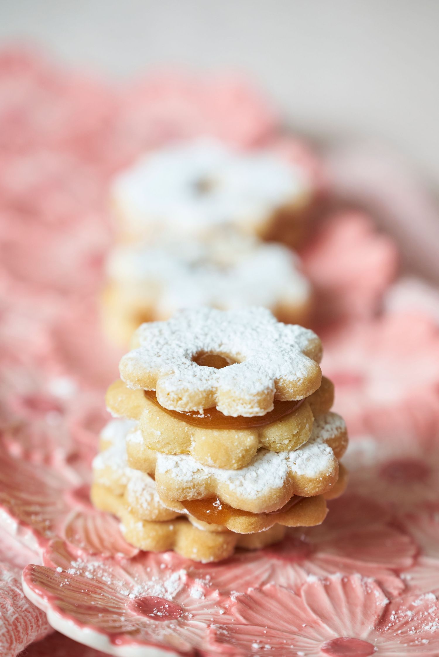 Delicate flower-shaped linzer cookies dusted with powdered sugar stacked on a pink glass plate.