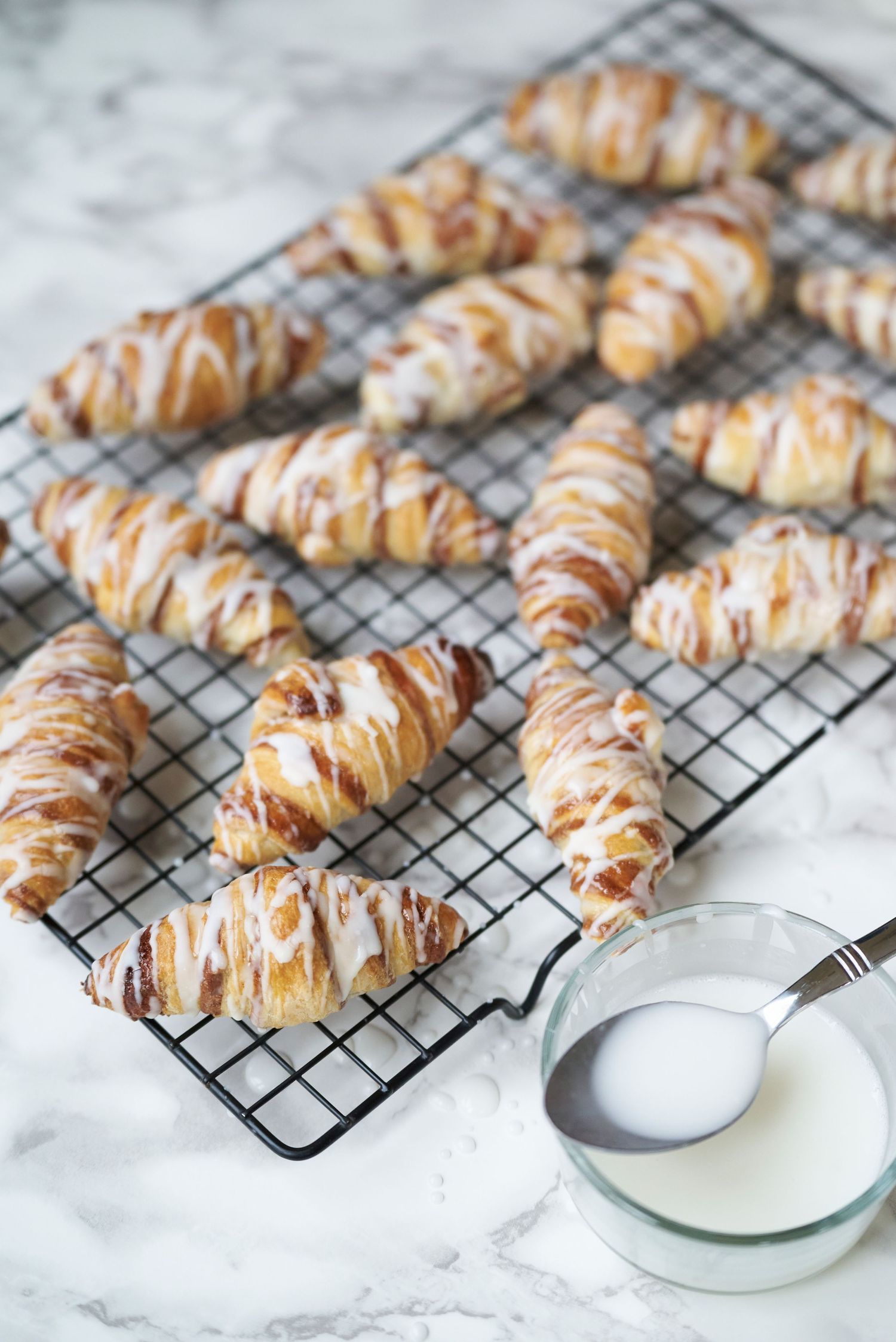 Freshly baked pastry twists drizzled with white icing cooling on a wire rack with glaze bowl nearby.